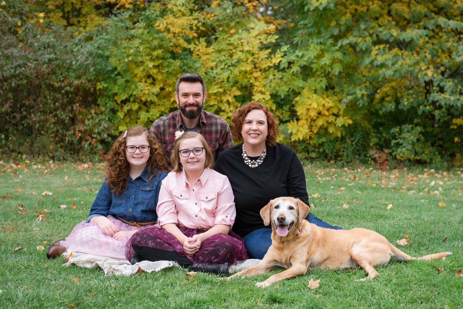 A golden retriever dog rests beside a group enjoying an autumn photo session on green grass with fall foliage.