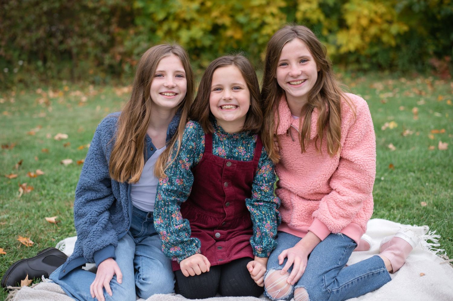 Three people sit close together on a blanket outdoors during autumn photo session with golden trees behind.