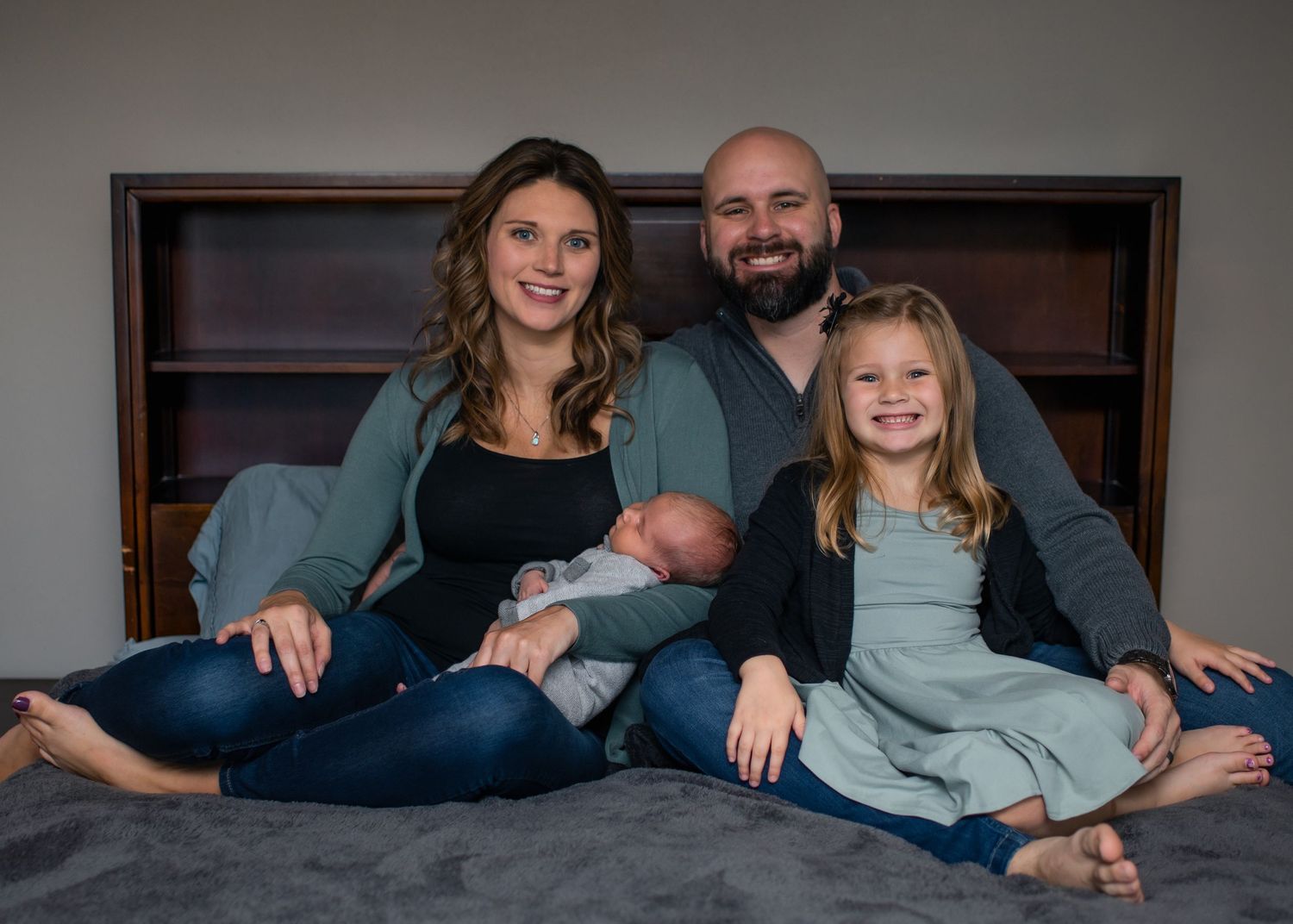 Family of four relaxes together on a gray bed with wooden headboard in casual indoor portrait setting.
