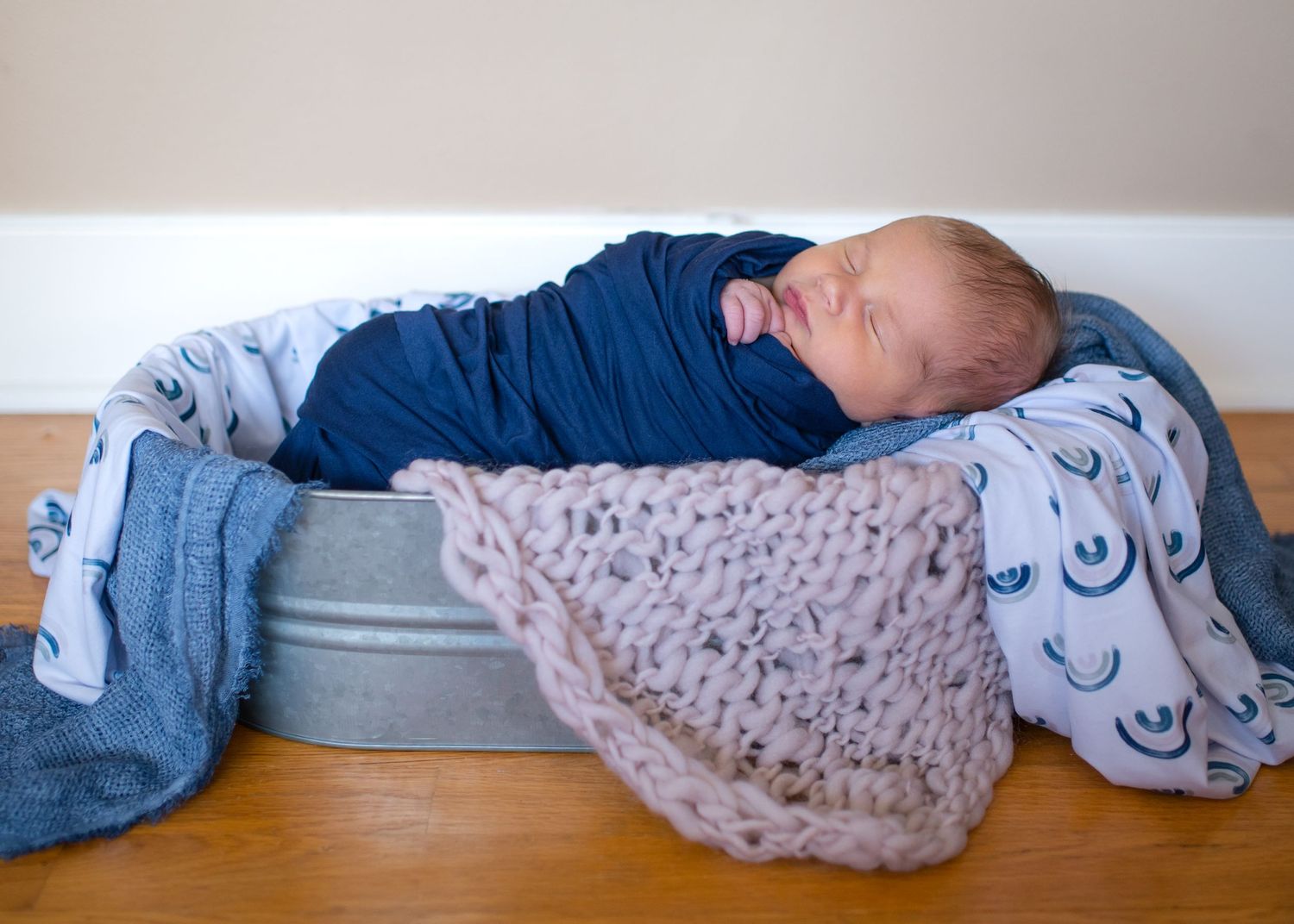 Sleeping newborn wrapped in blue swaddle on metal basin with knit blanket.