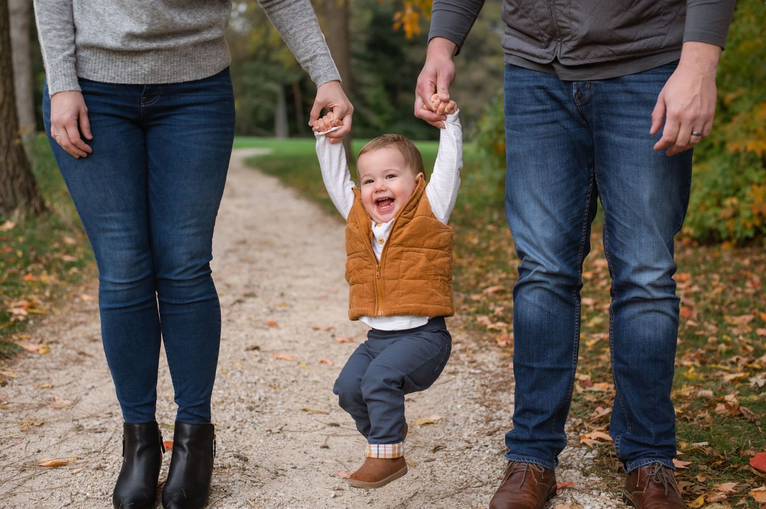 Small child in brown vest swings joyfully between two adults on an autumn path.