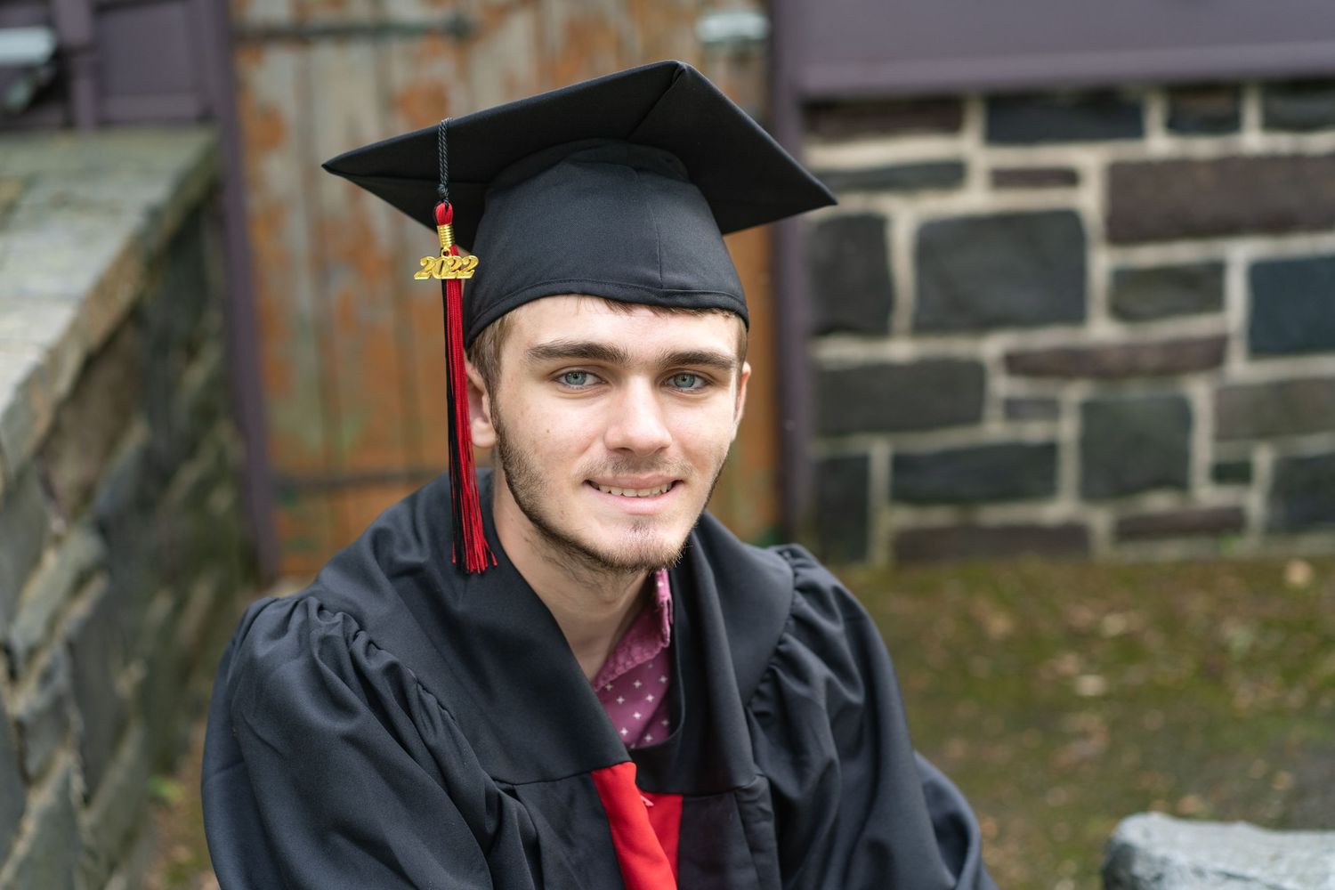 Graduate wearing black cap and gown with red tassel against stone wall.