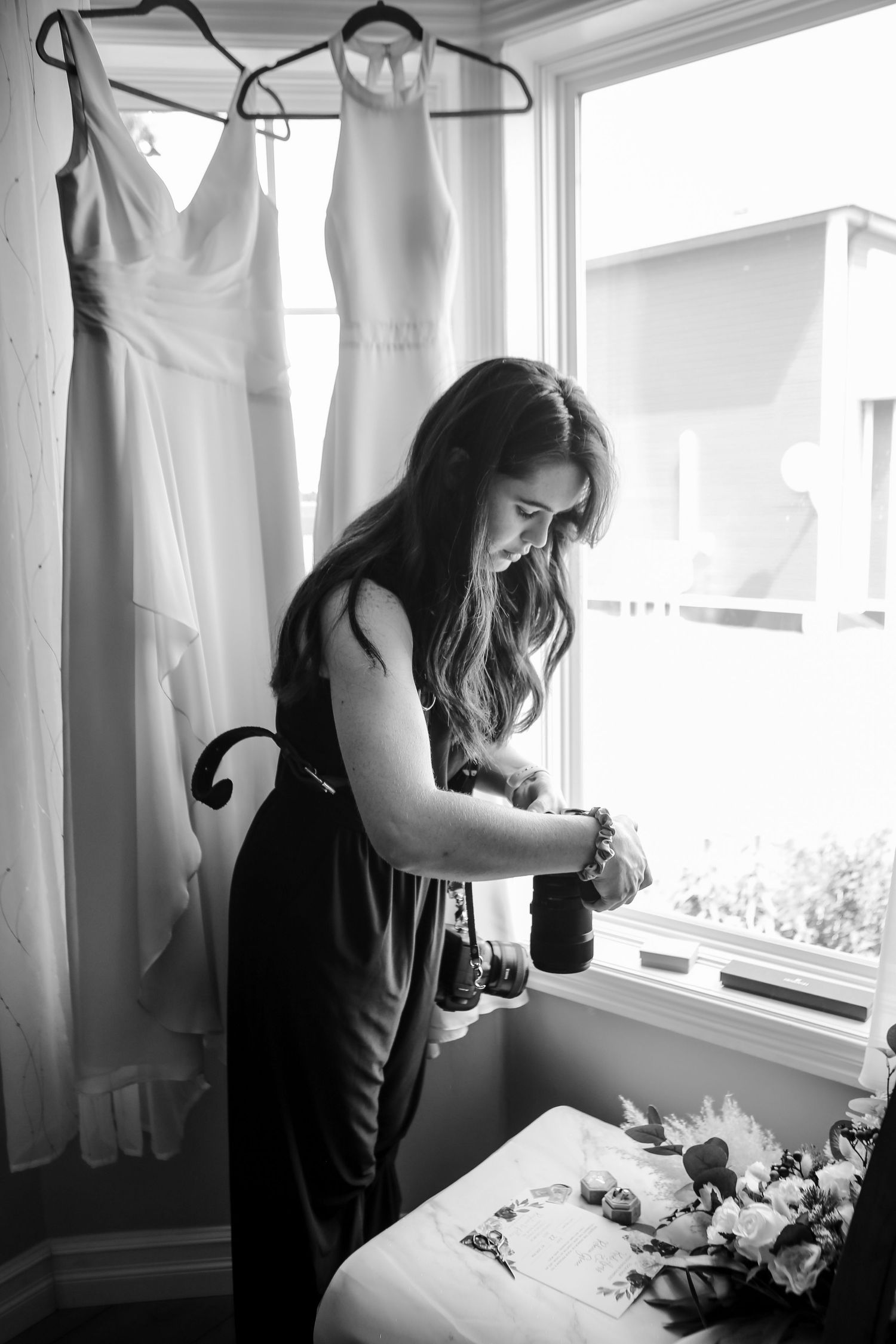 Black and white image showing wedding dress preparations with dresses hanging in window light.