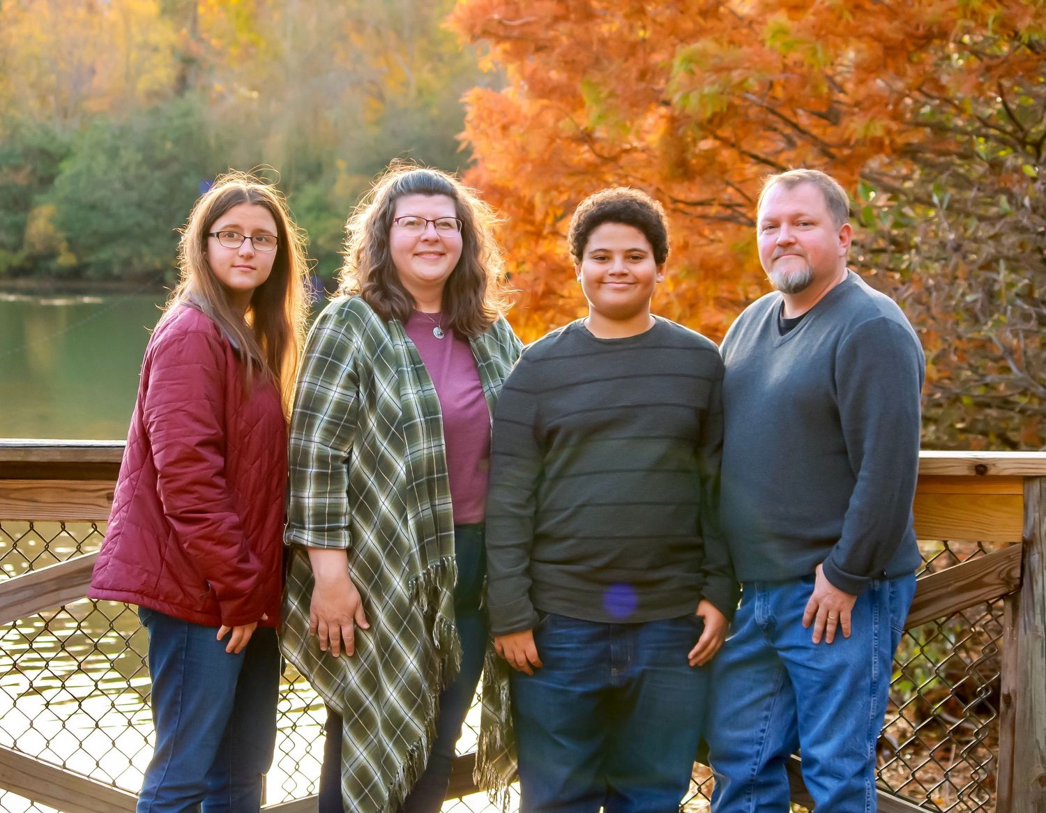 A group of four people standing together on a deck with autumn foliage and a lake in the background.