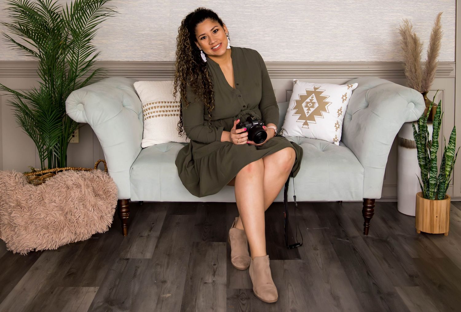 Woman in olive dress sits on gray couch with camera in modern boho-styled living room.