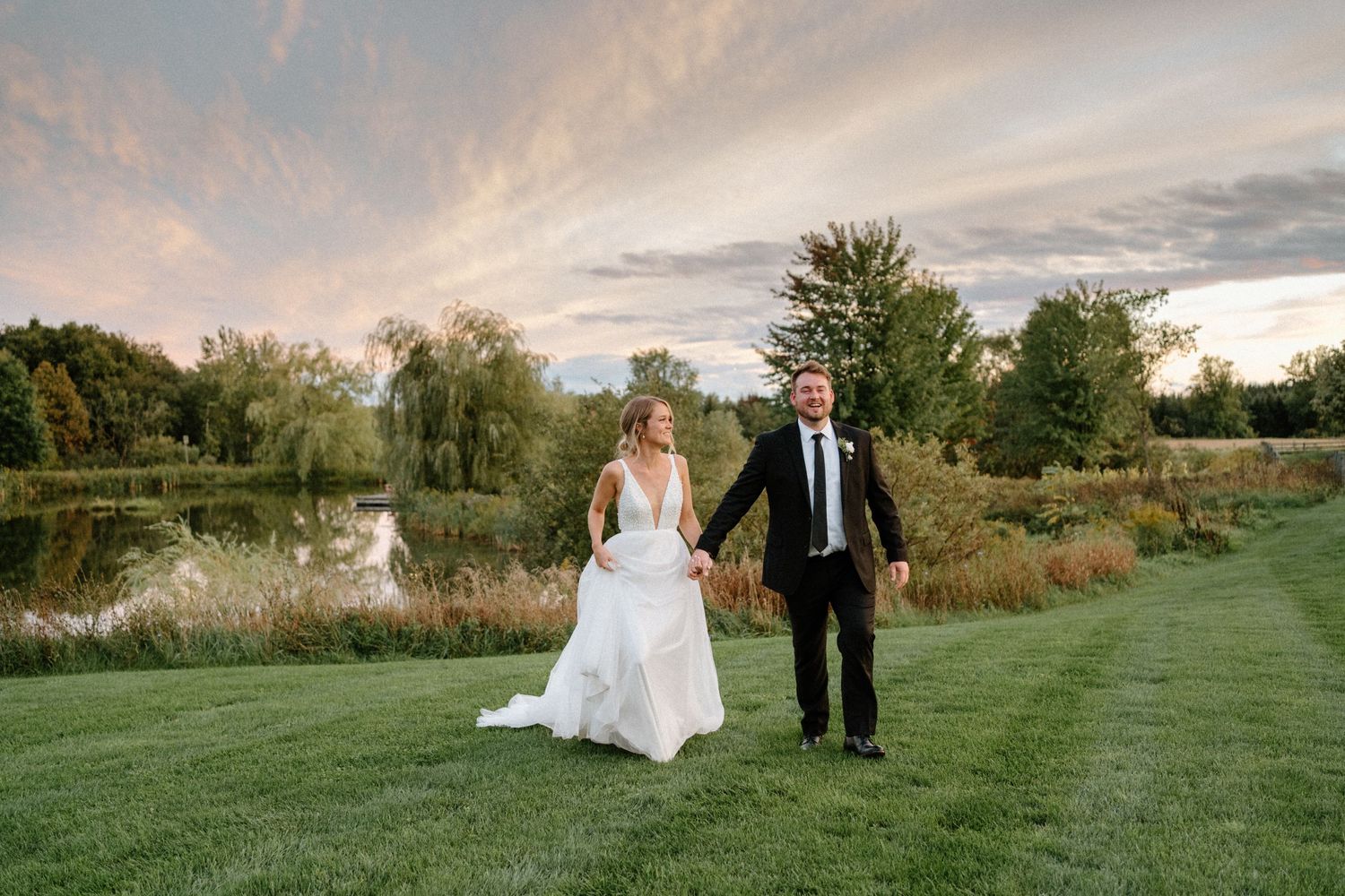 A wedding couple walks hand in hand across a manicured lawn at sunset.