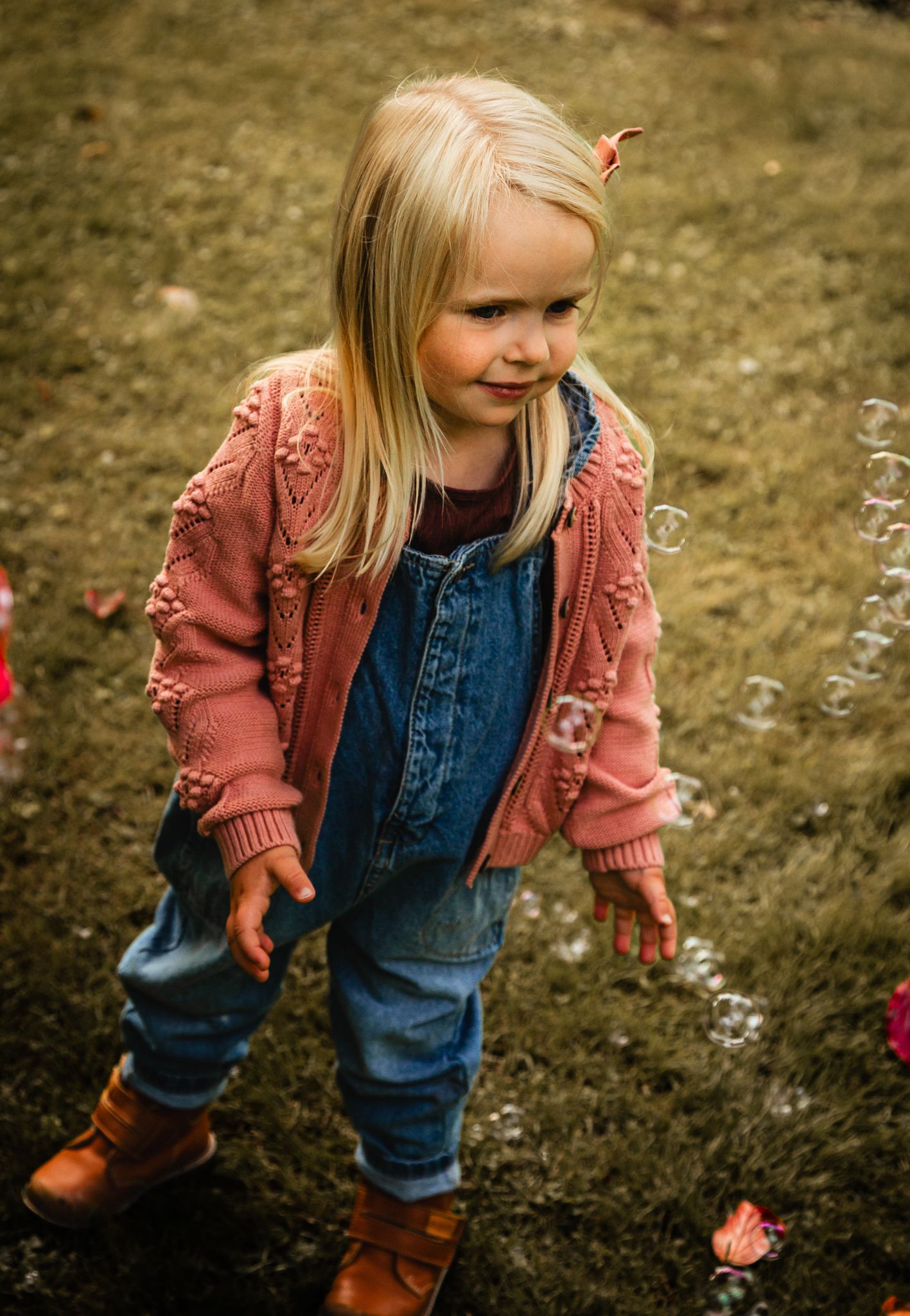 A toddler in denim overalls and pink jacket plays outdoors.