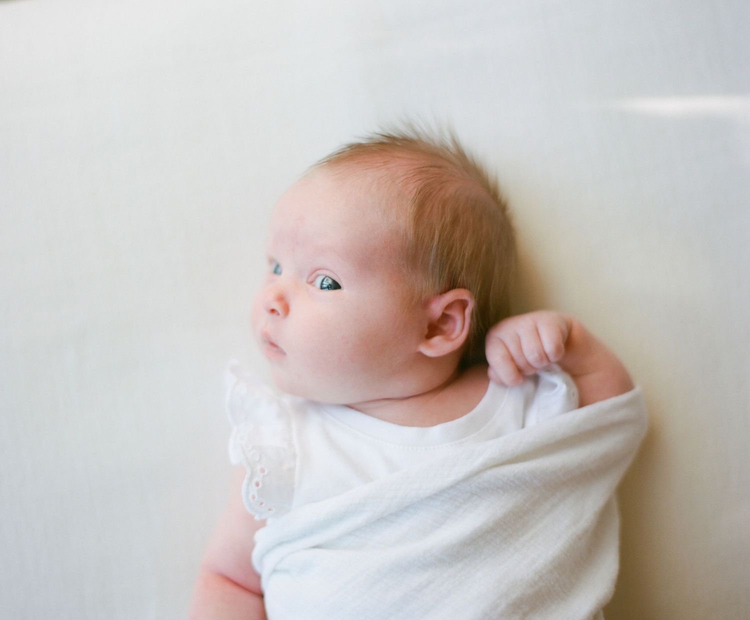 3 month baby girl in white lace capped onesie with blue eyes looking at camera slightly out of corner of eyes on a white
