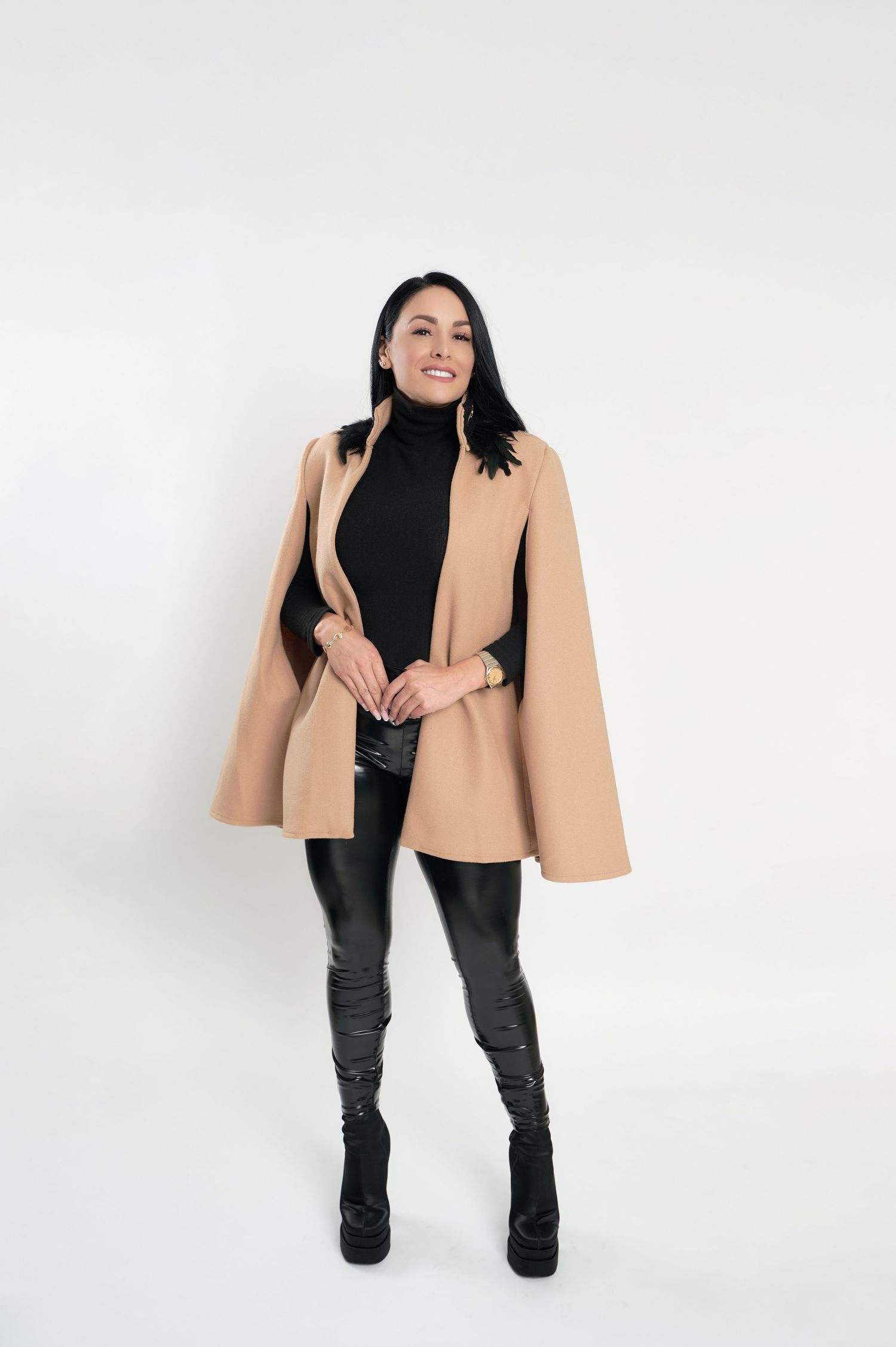 Fashion model poses in a camel cape coat, black turtleneck and leather pants against white background.
