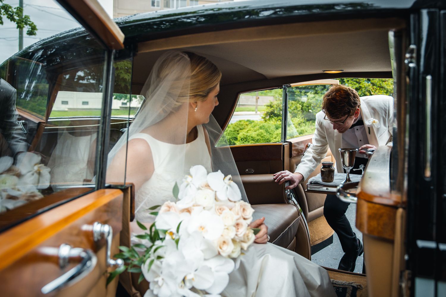 A newlywed couple sharing a tender moment in a classic Rolls Royce car. The bride, with her veil and bouquet, sits grace