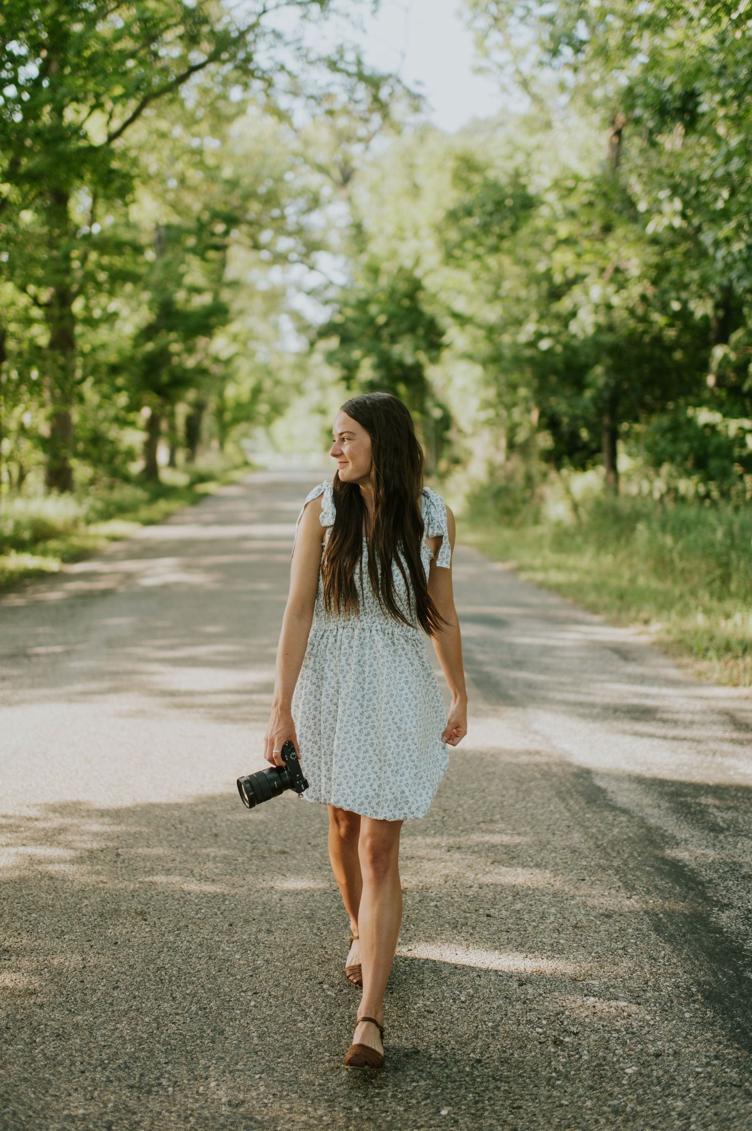 Person in a light summer dress walking down a tree-lined path holding a camera.