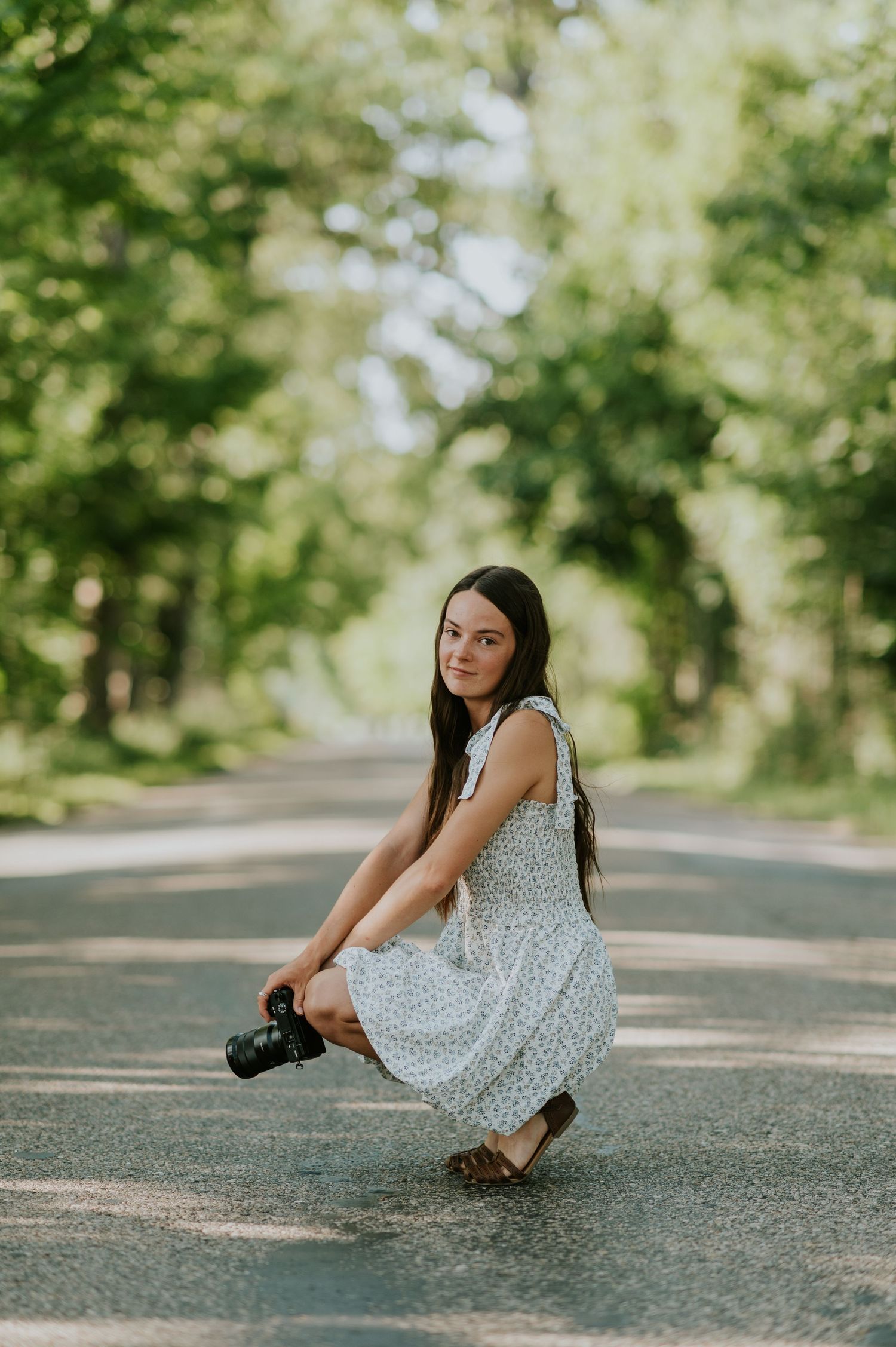 Person in a floral dress crouching on a tree-lined path with a camera.