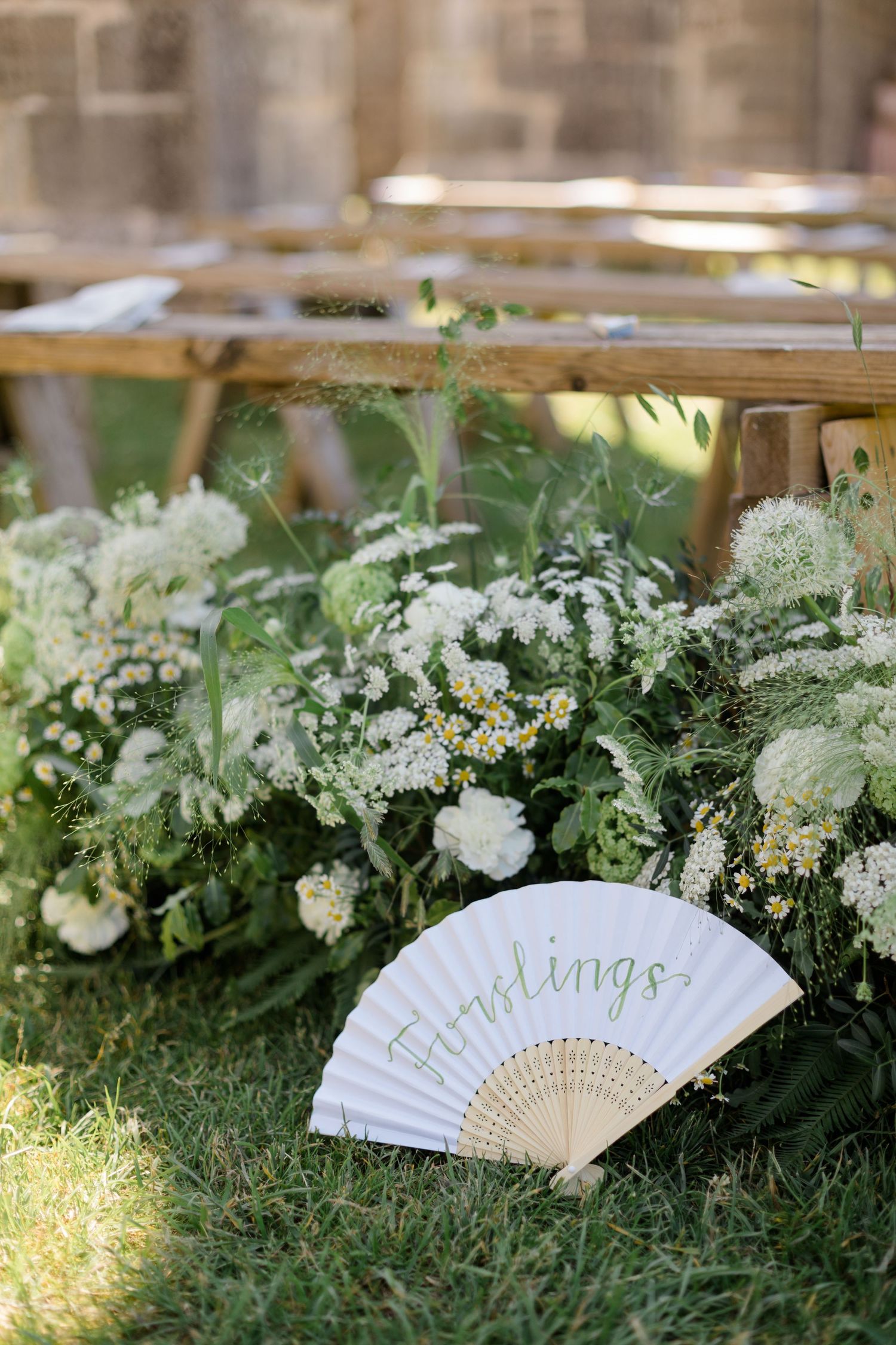 Decorative white hand fan nestled among lush green and white floral arrangements outdoors.