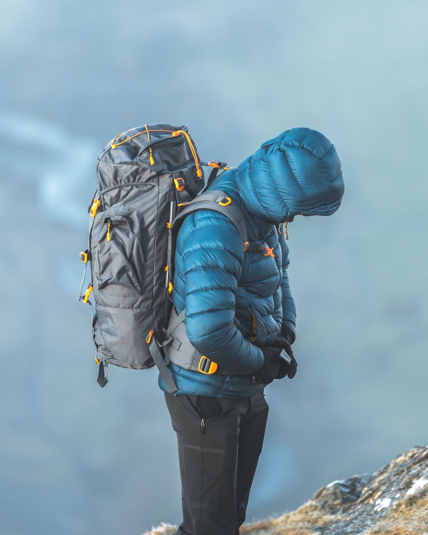 Hiker wearing a blue down jacket carries two large backpacks against a misty mountain backdrop.