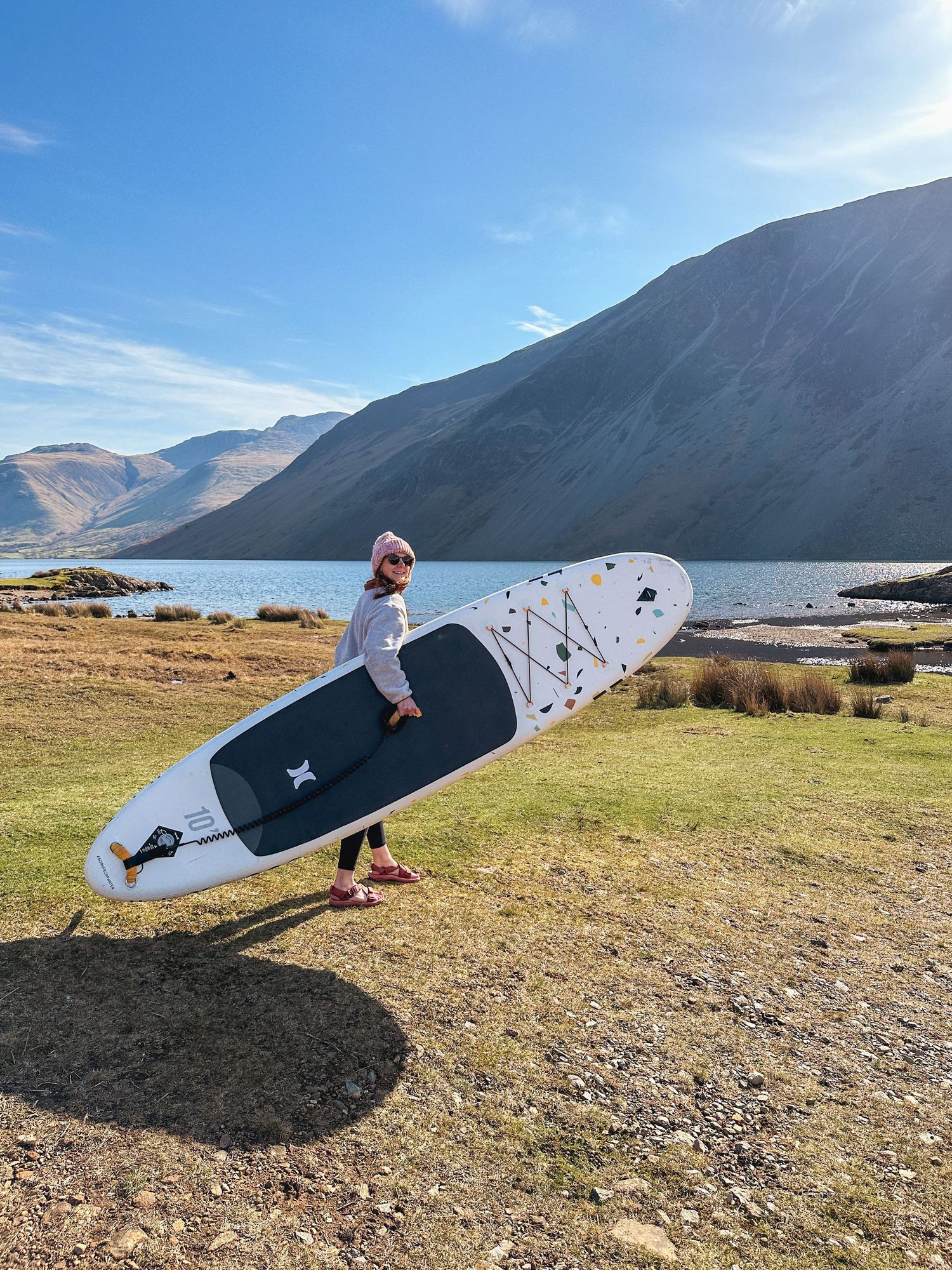 Person carrying a paddleboard beside a scenic mountain lake under blue skies.