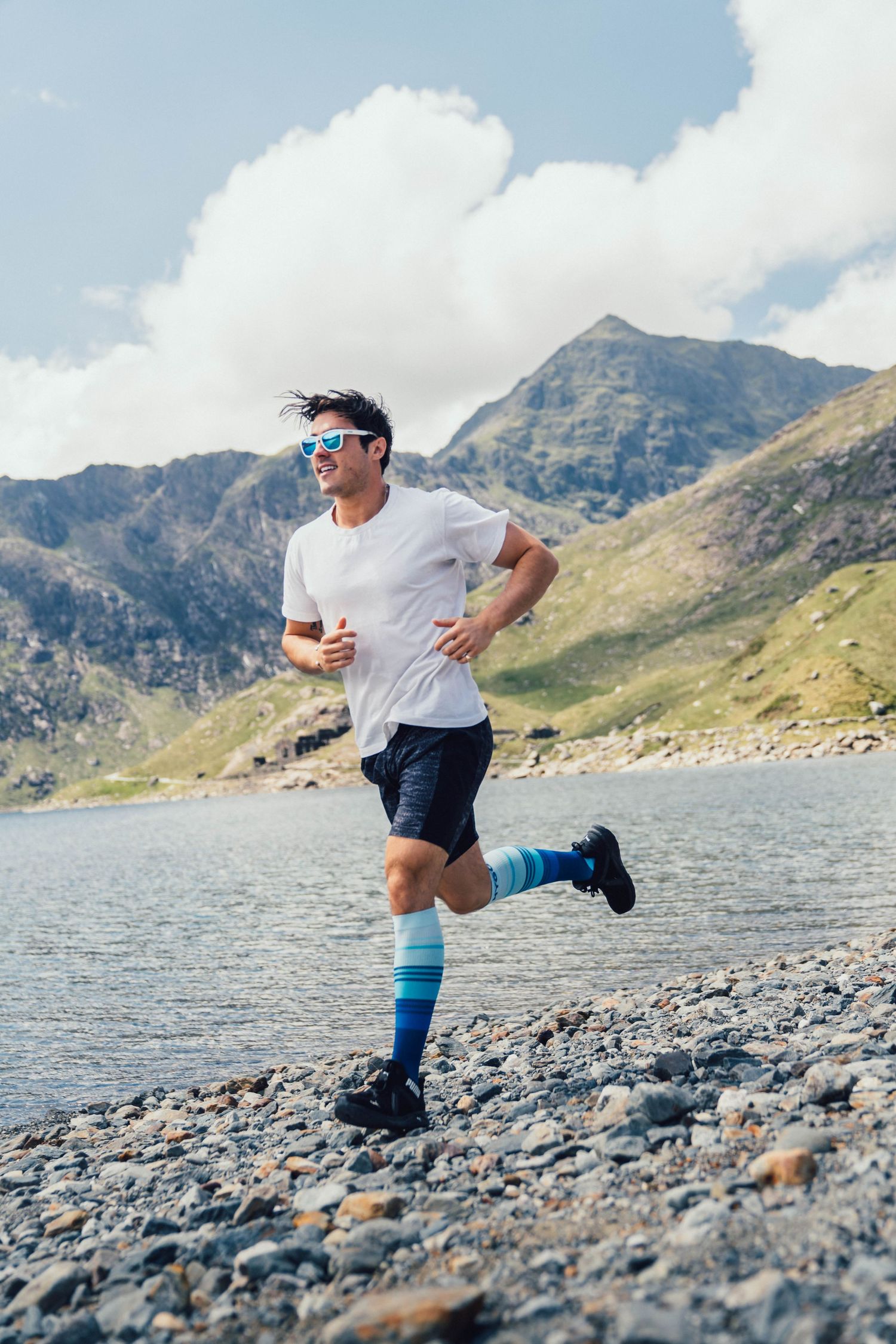 Runner in white shirt and blue socks on rocky mountain trail near lake.