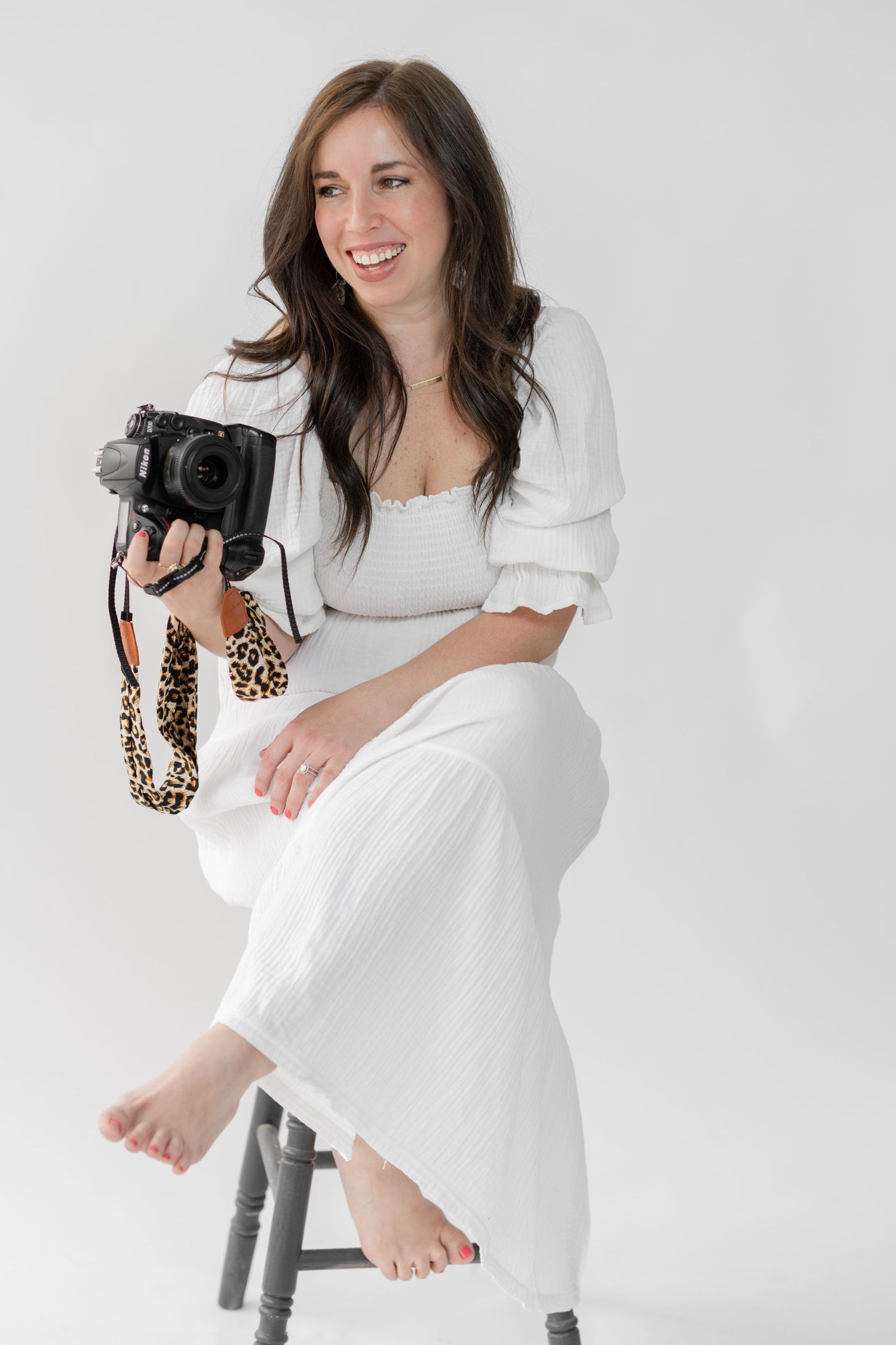 headshot of ashley line sitting on a bench holding her camera.  laughing over her shoulder in a white dress