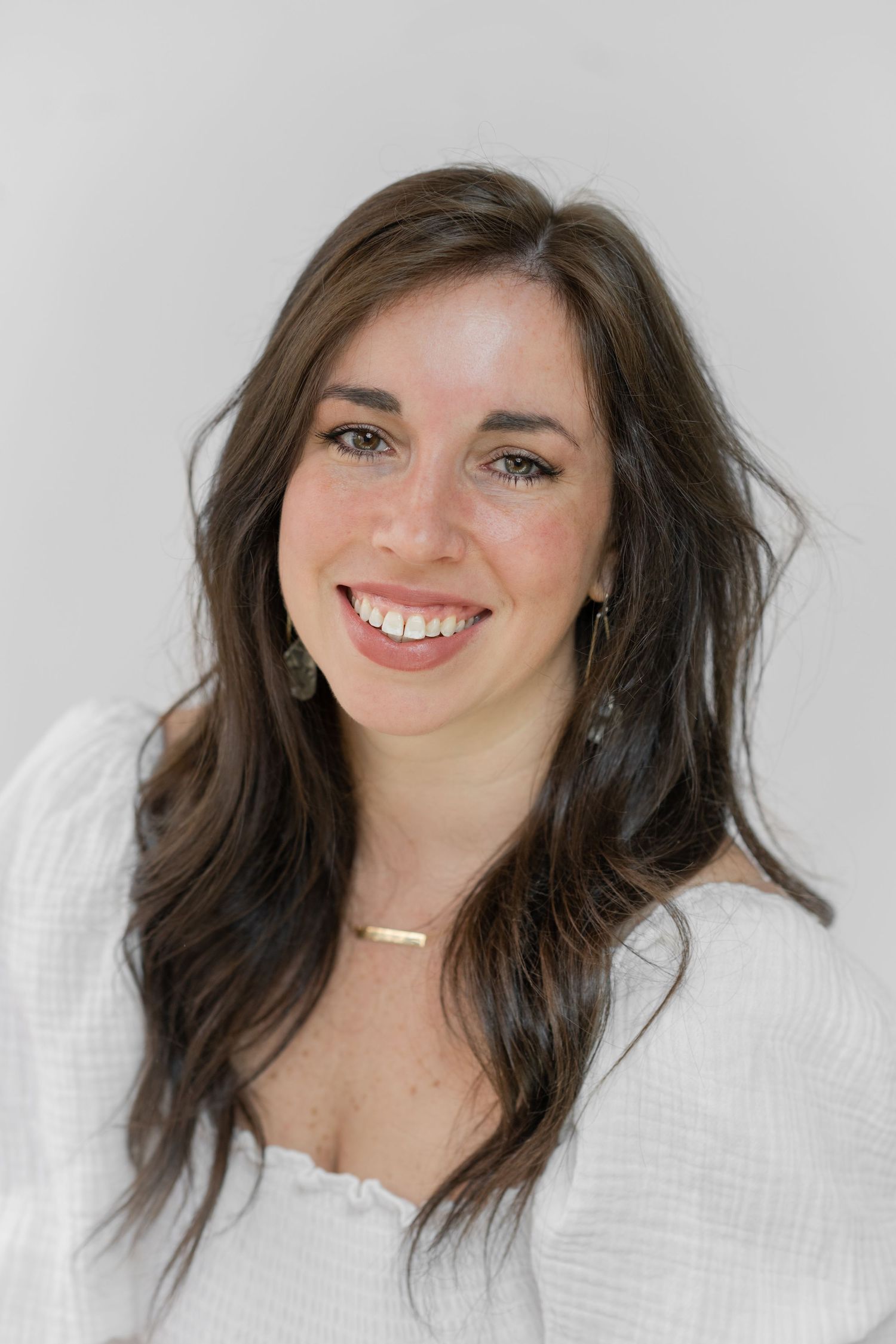headshot of ashley line photography smiling at the camera.  dark curly hair white dress and gold necklace