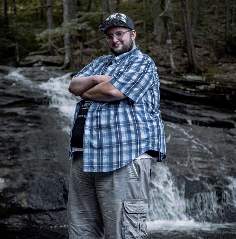 A person in a plaid shirt and baseball cap poses with crossed arms in front of a cascading forest waterfall.