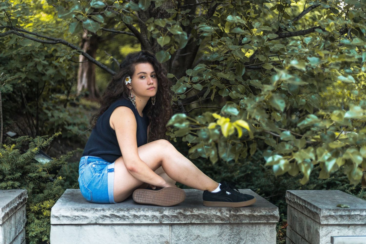 Person in blue shorts and black top sitting on concrete wall surrounded by leafy green foliage in garden setting.