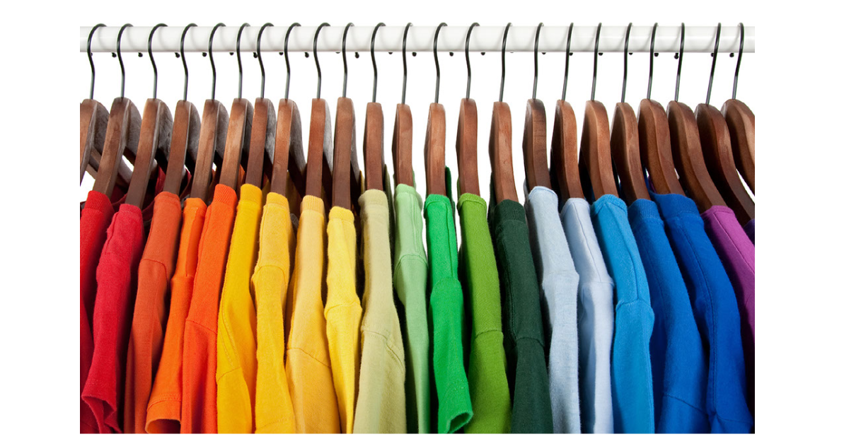 Rainbow collection of shirts arranged by color on hangers in a closet.