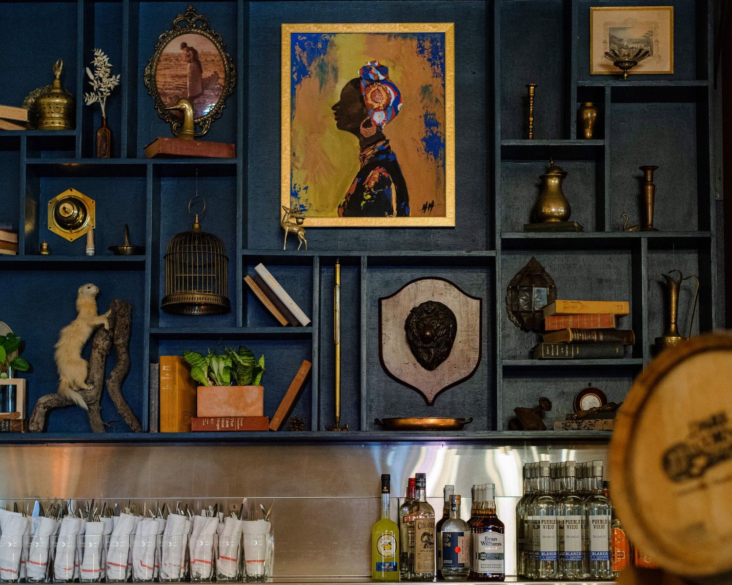 Stylish bar shelf display with vintage decor, artwork, and bottles arranged on blue wooden compartments.