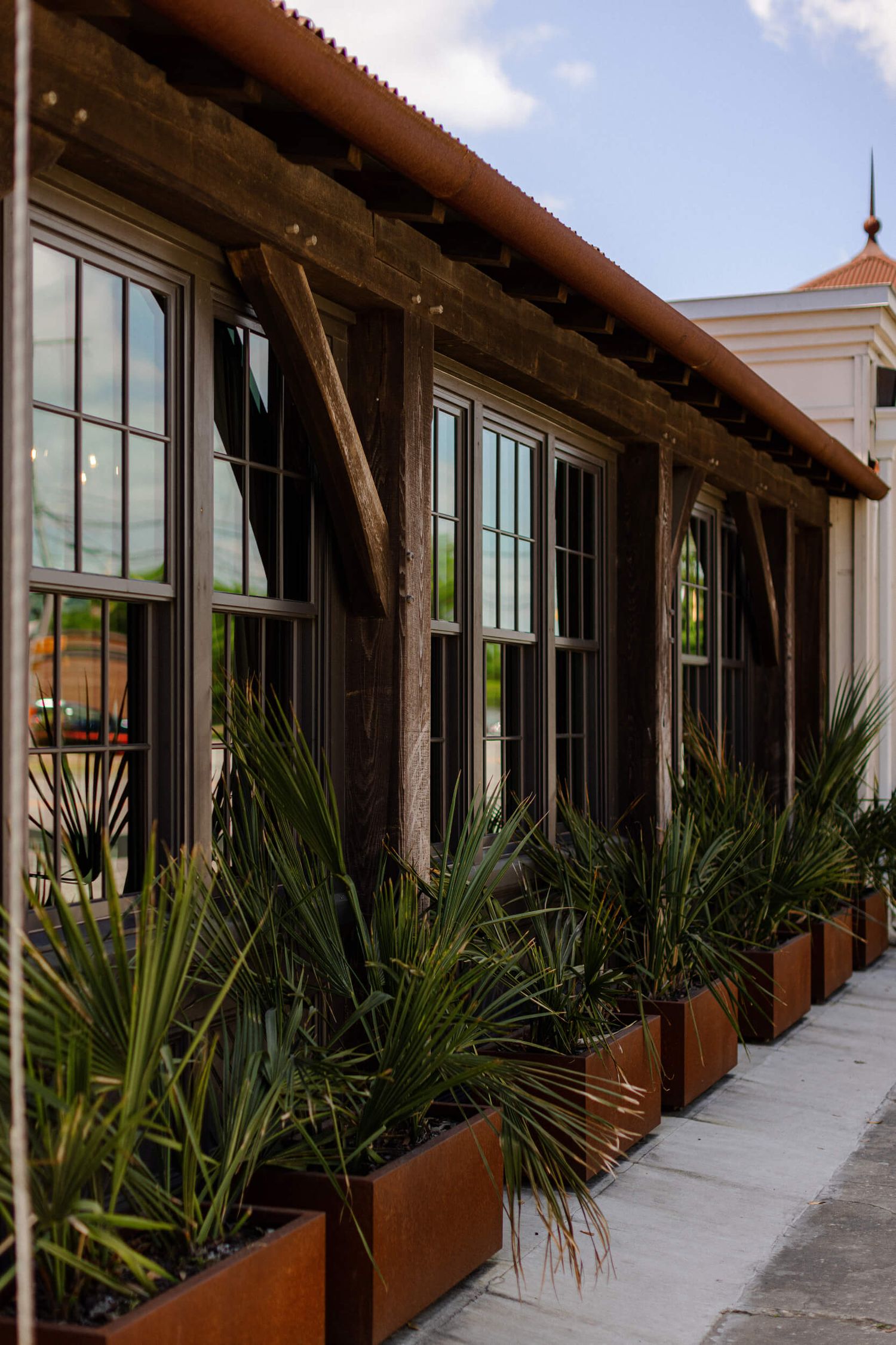 Modern restaurant exterior with dark wooden beams and planted palm grasses in metal planters.
