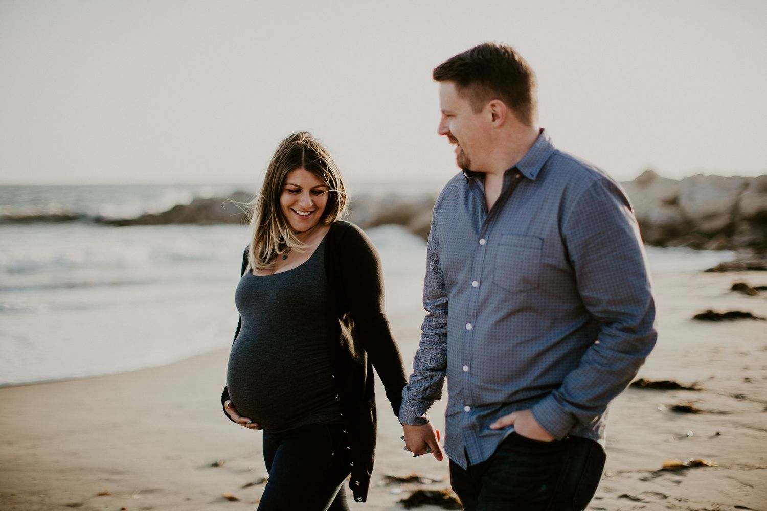 Couple takes romantic sunset stroll along rocky beach during maternity photoshoot.