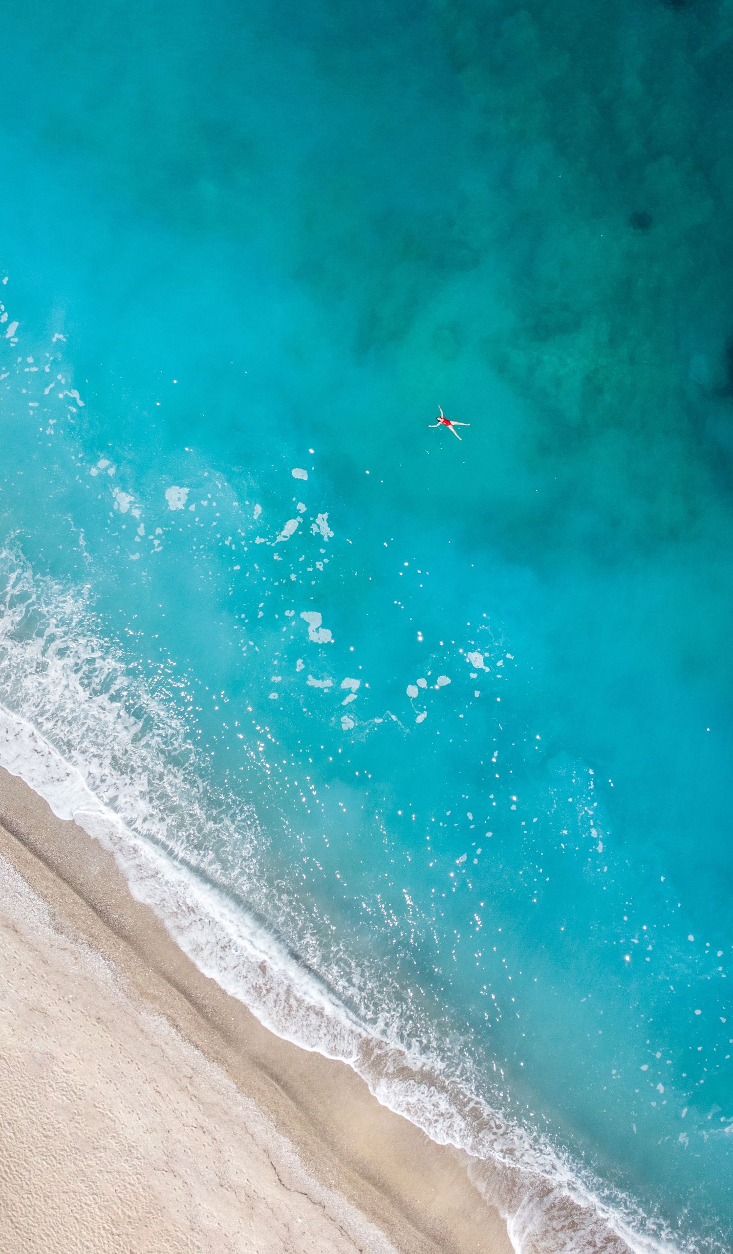 Aerial drone view of a lone swimmer in stunning turquoise ocean waters meeting a sandy beach shoreline.