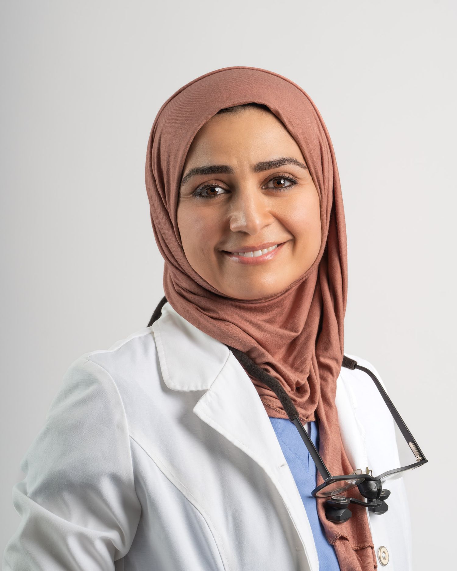 Professional headshot of a healthcare worker wearing hijab and white coat with stethoscope.