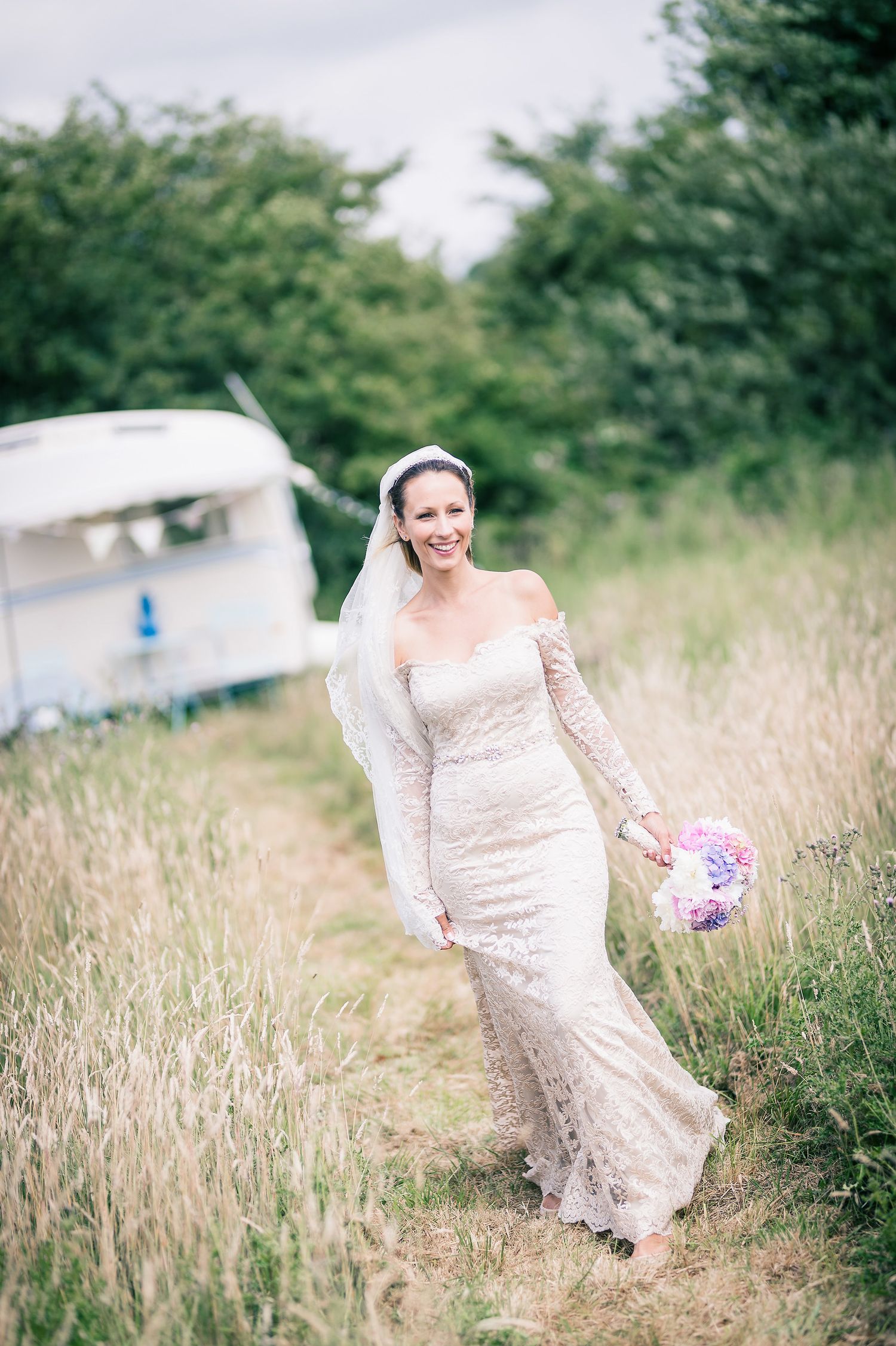 A bride in a fitted lace gown walks through tall grass carrying a pink bouquet near a vintage trailer.