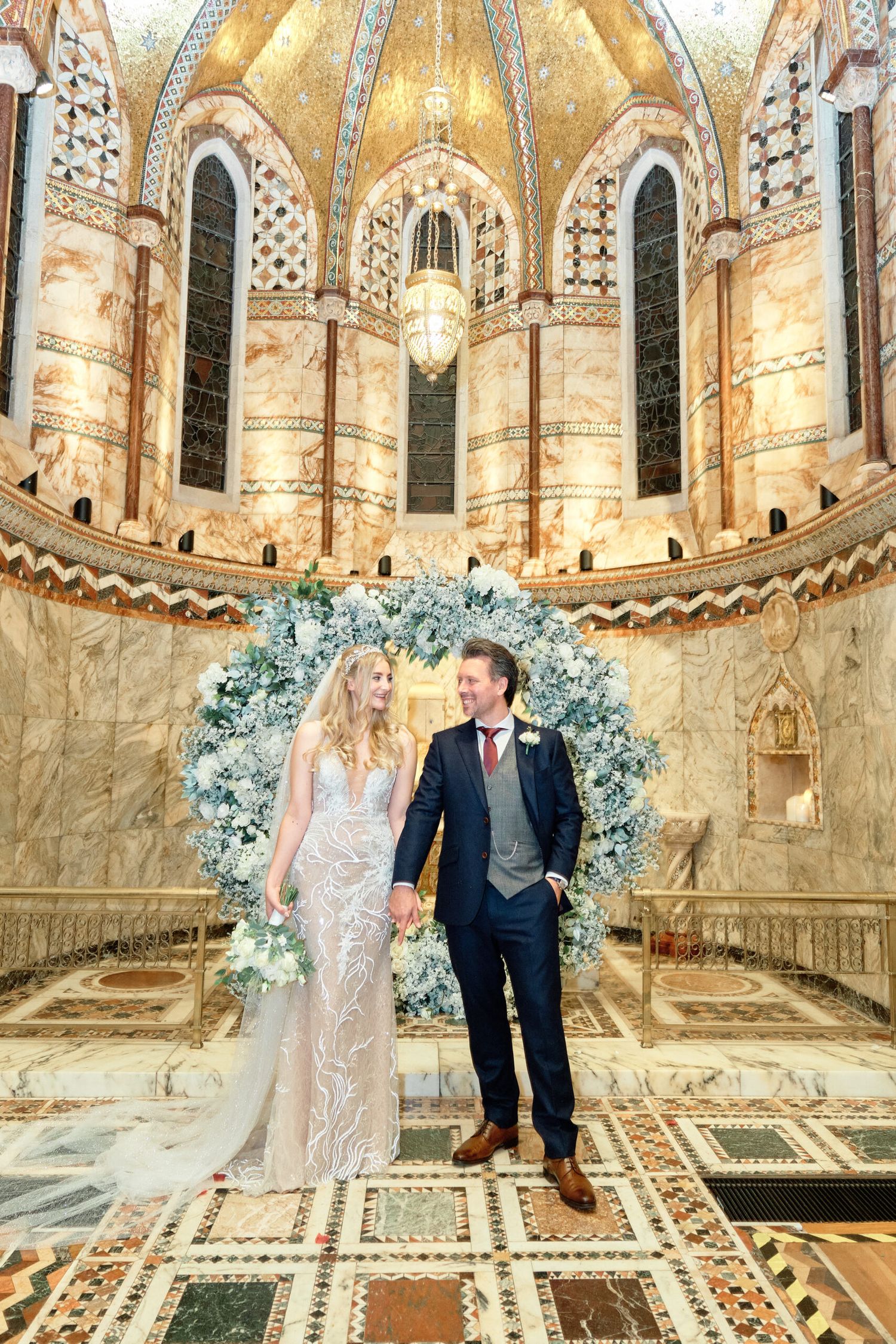 A wedding couple stands under an ornate floral arch in a grand cathedral with golden domed ceiling.