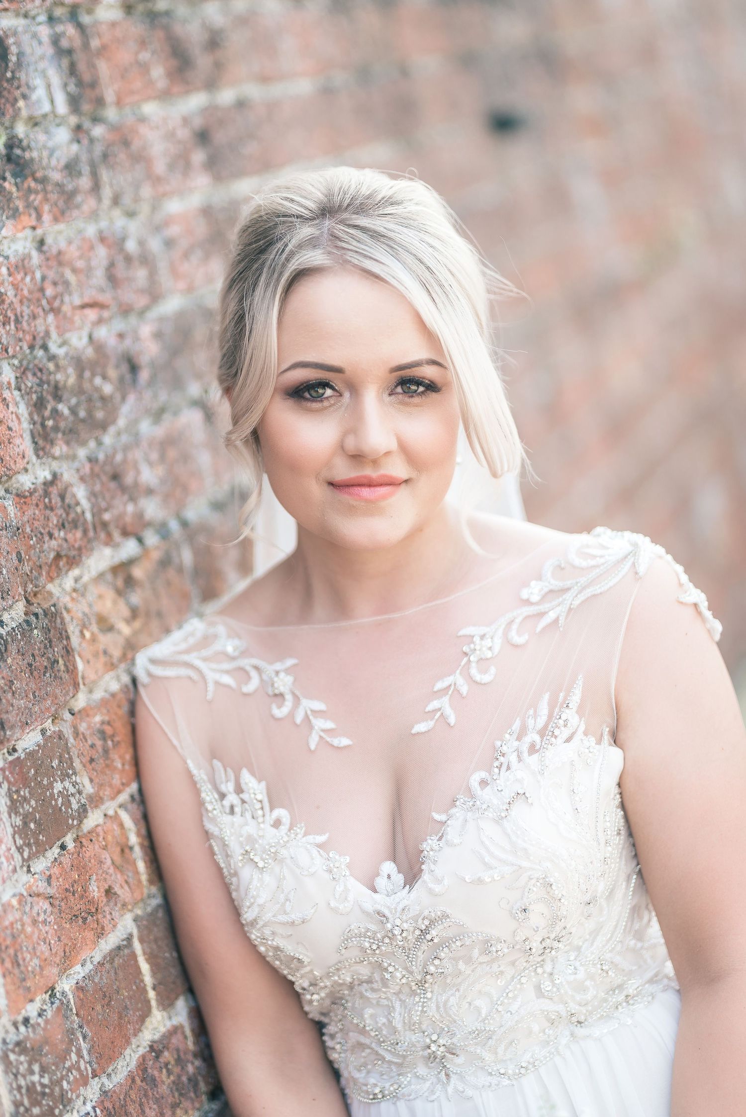 A portrait of a bride wearing an elegant lace-detailed dress against a brick wall background.