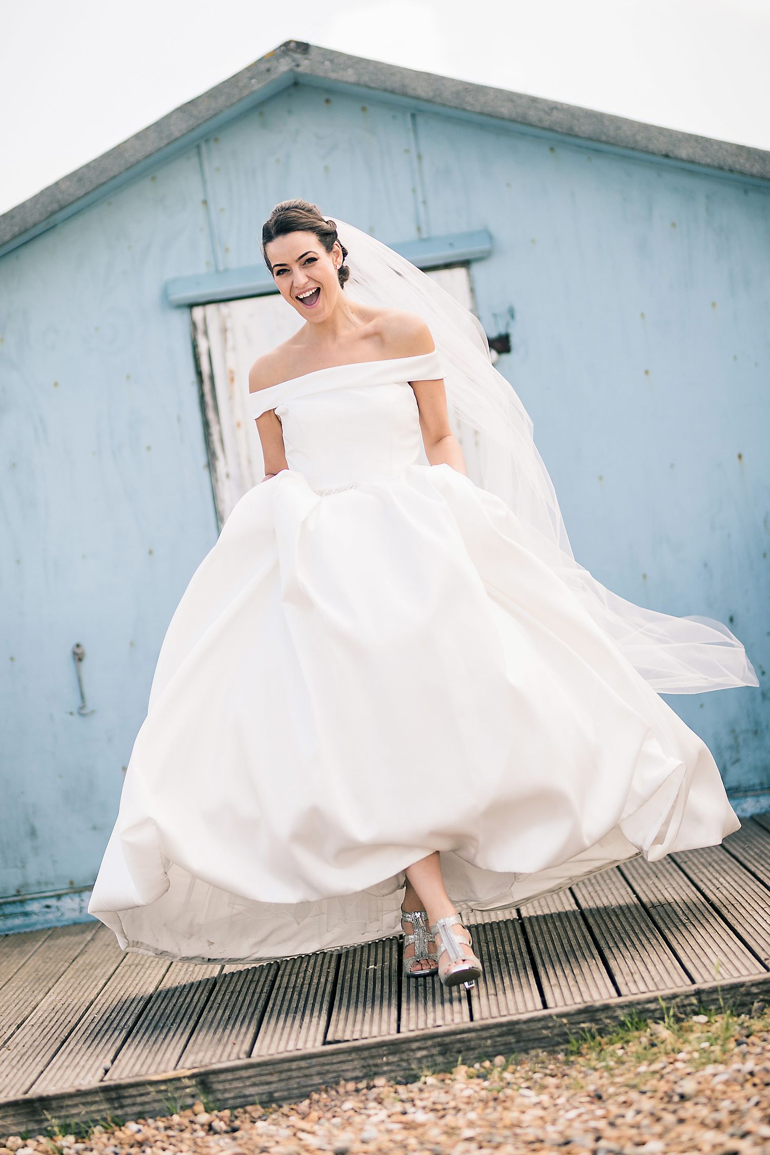 A bride in an off-shoulder ballgown twirls on a wooden deck in front of a blue beach house.