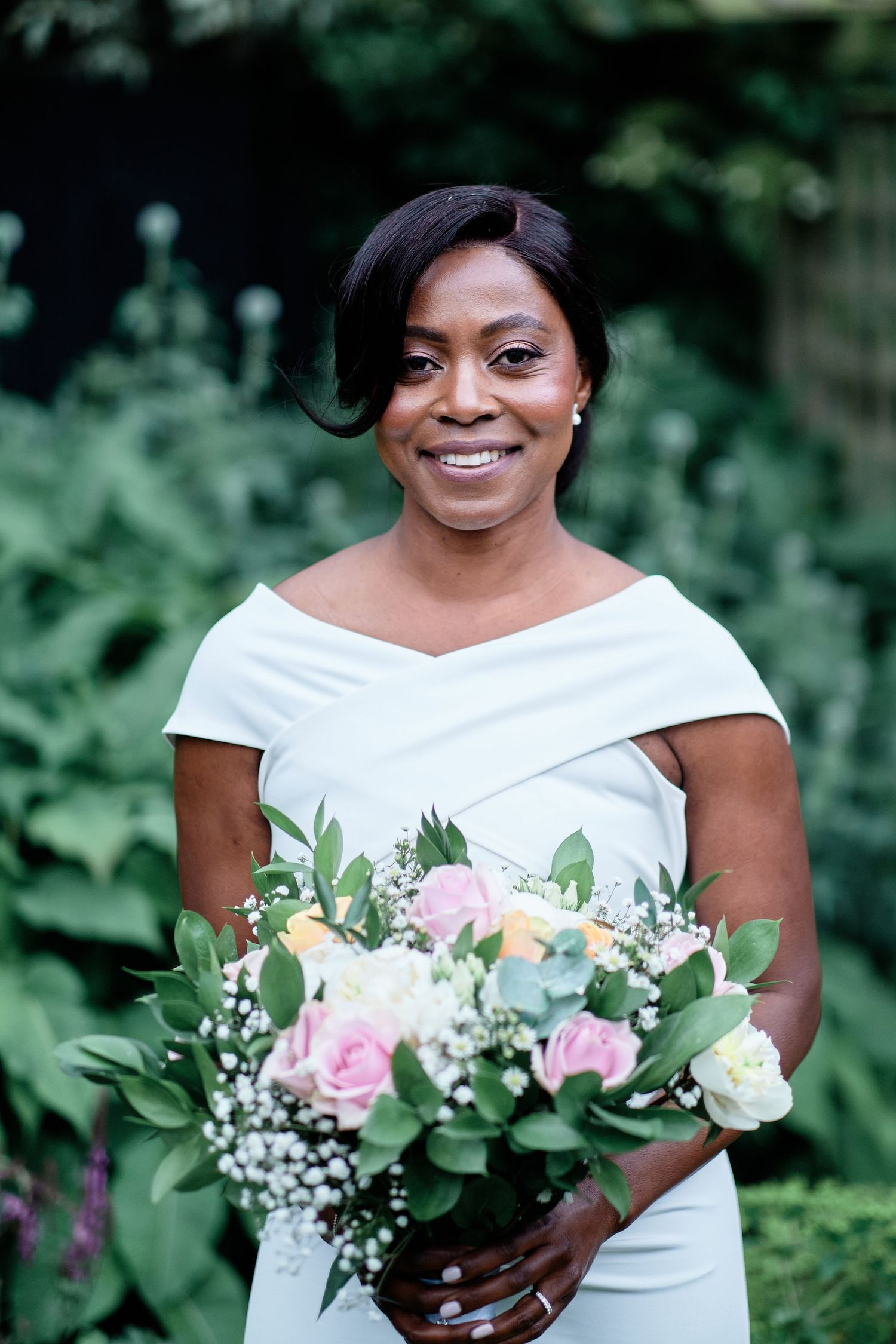 A radiant bride in an elegant off-shoulder white dress holds a colorful bouquet against a garden backdrop.