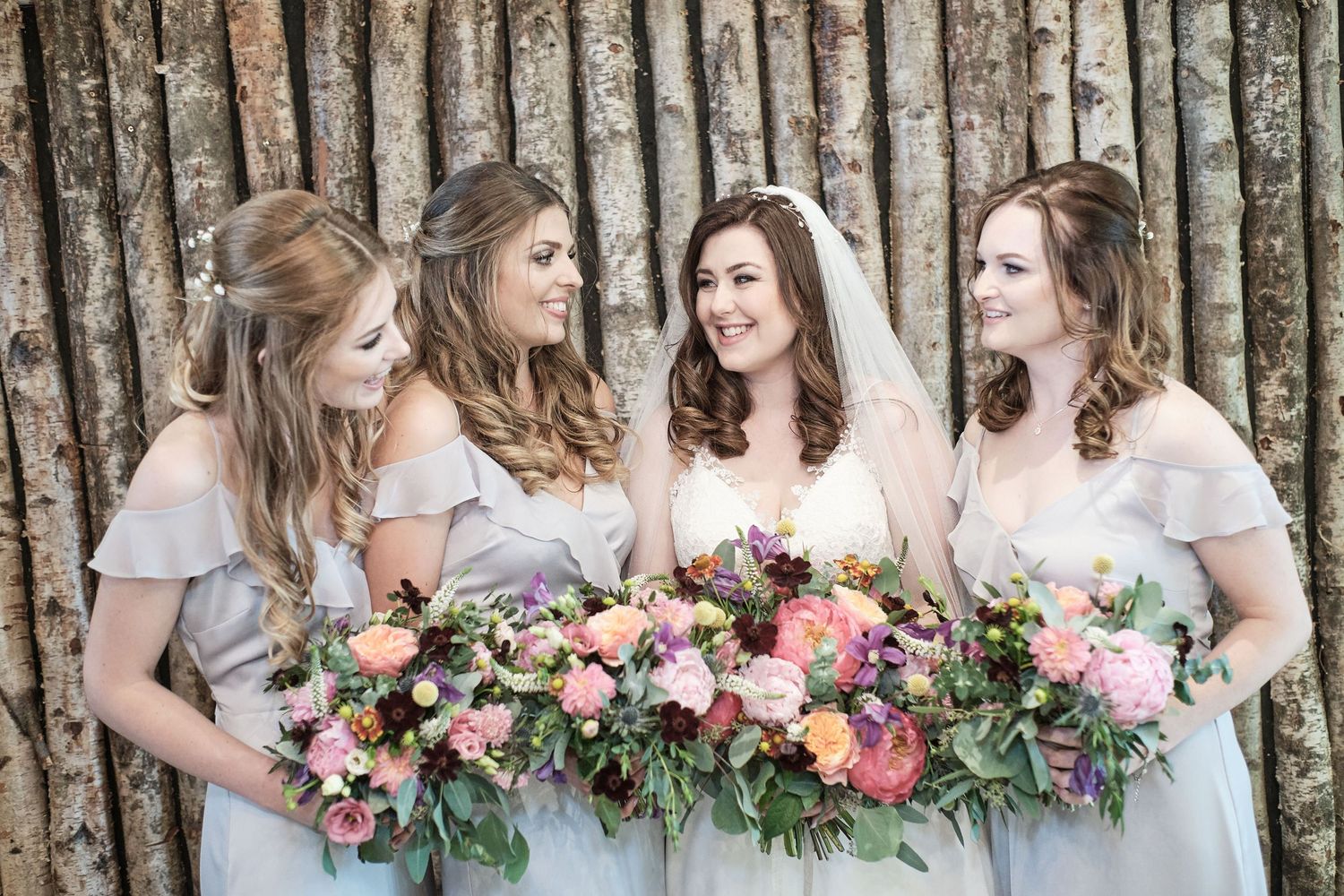 Four bridesmaids in light blue dresses hold colorful floral bouquets against a rustic backdrop.