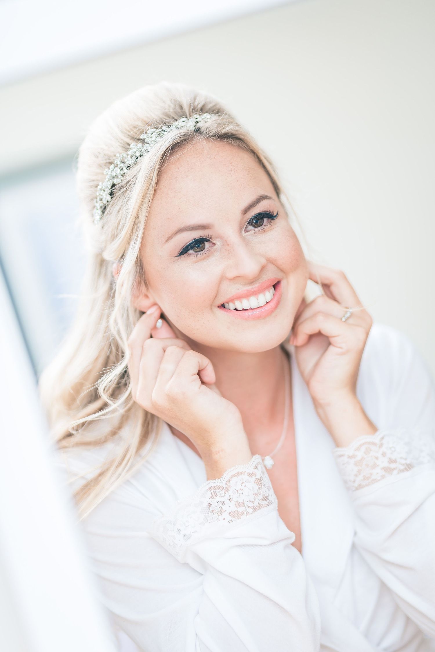 Bride wearing lace robe and crystal headpiece smiles during wedding preparations.
