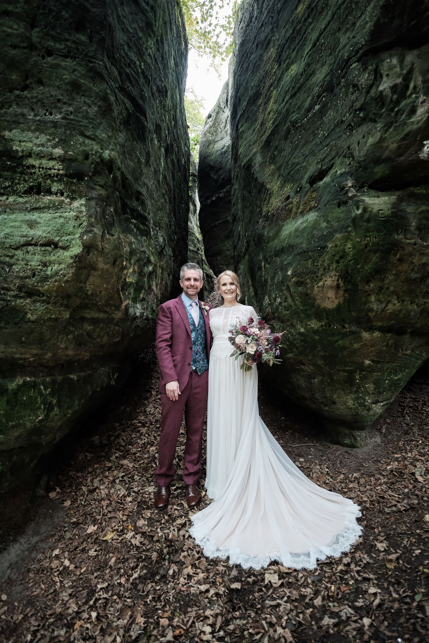 Wedding couple poses between dramatic mossy rock formations in an enchanting woodland setting.