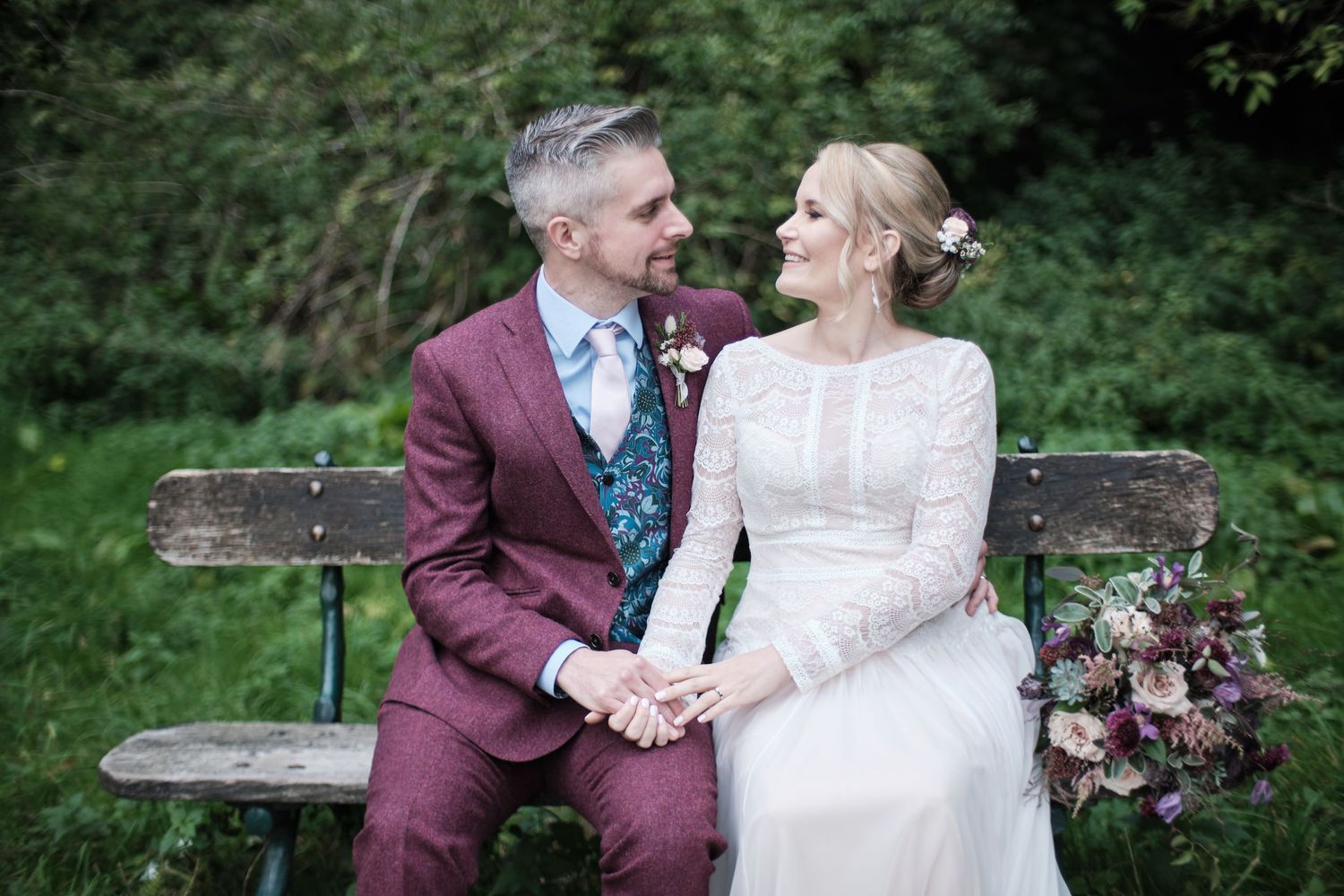 A wedding couple shares a tender moment while sitting on a wooden bench in a garden setting.