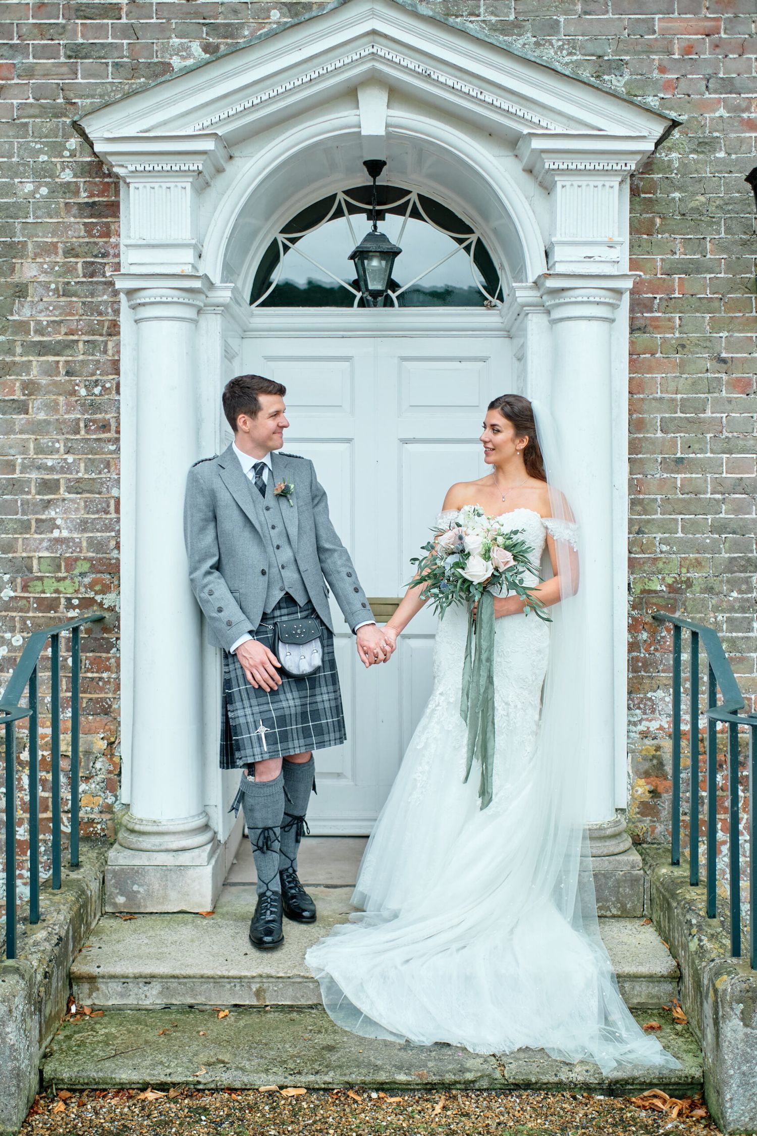 Bride in flowing gown and groom in traditional kilt hold hands at elegant white doorway entrance.