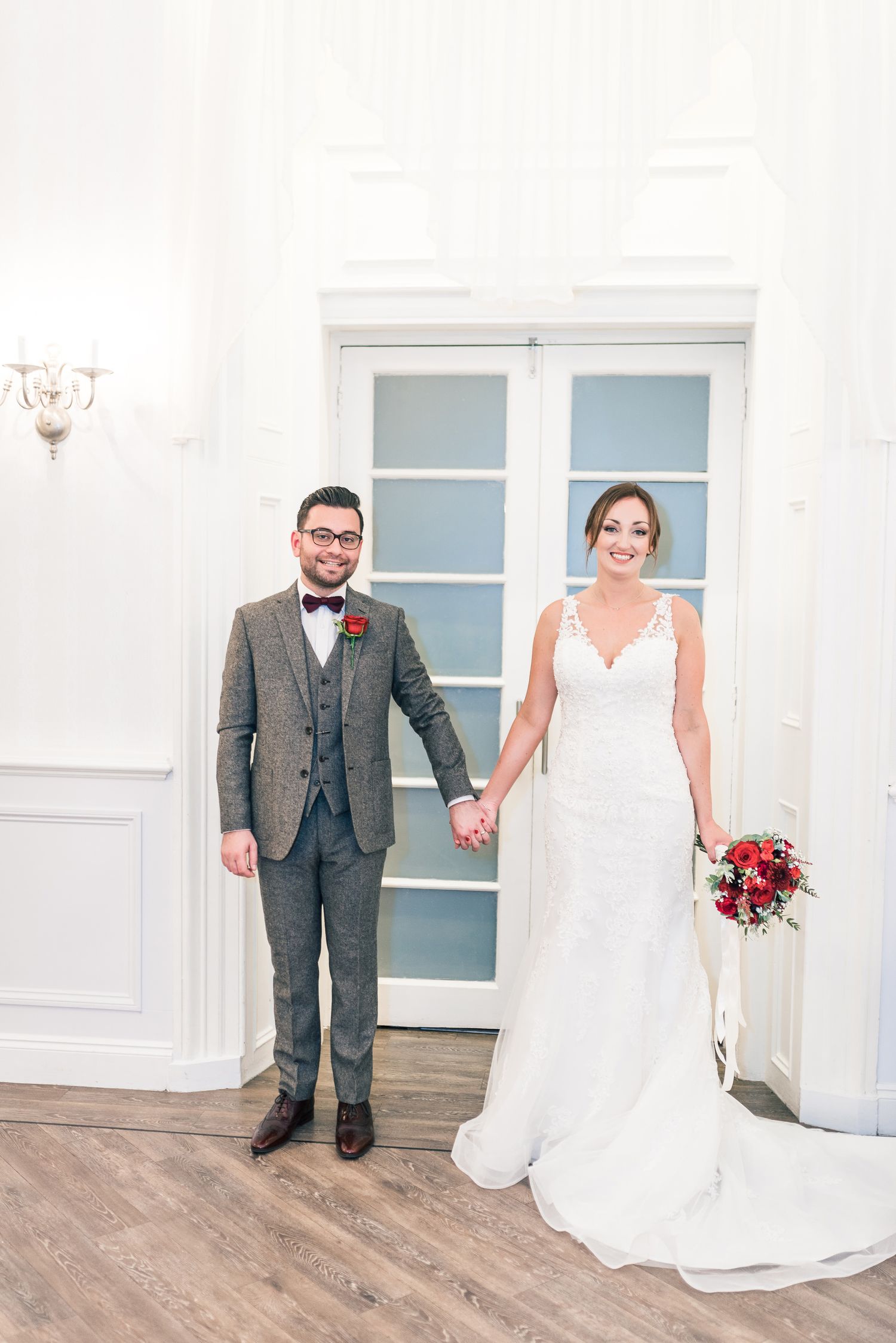 A wedding couple poses in formal attire against a white wall with french doors.