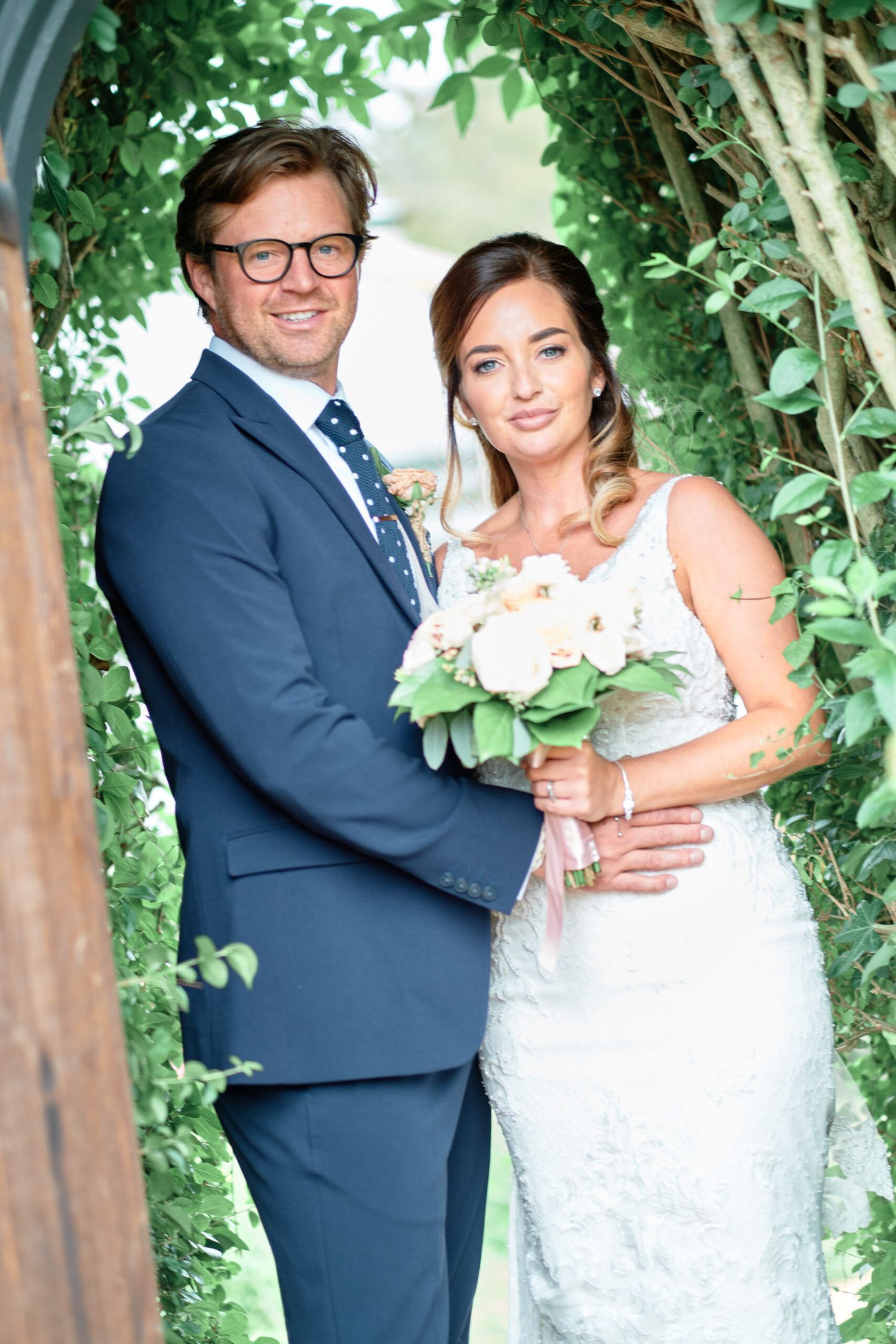 Couple poses under green foliage arch on wedding day with white floral bouquet.