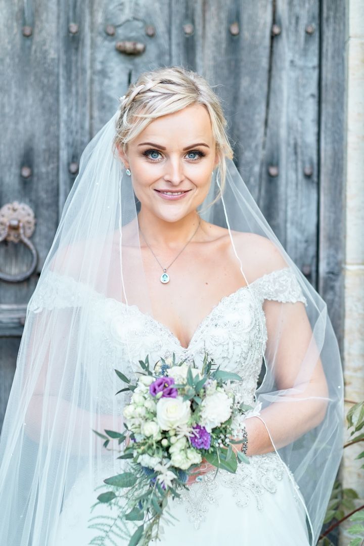 A bride in a lace wedding dress with veil poses against a rustic wooden door while holding a purple and white bouquet.