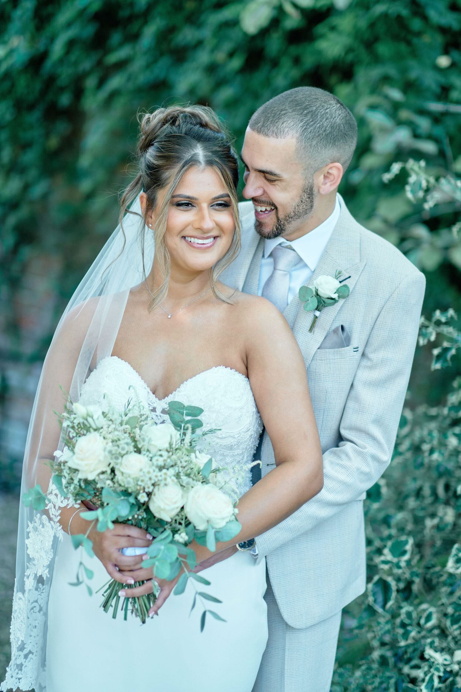 A joyful wedding couple in light colored attire embrace while holding a white floral bouquet outdoors.