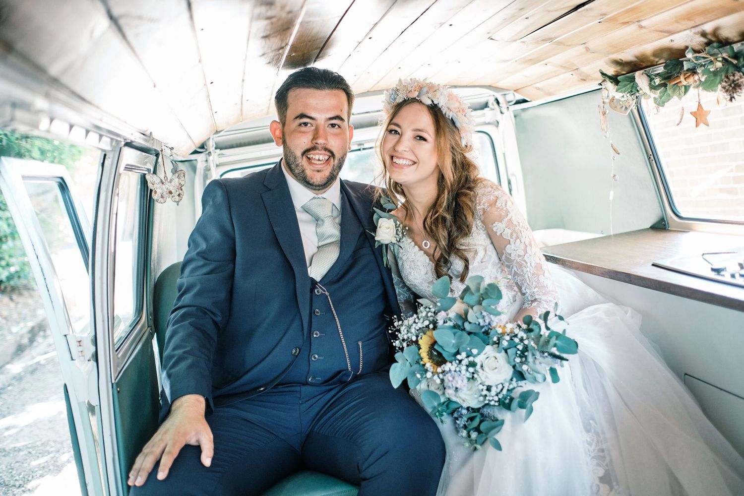 A wedding couple sits inside a vintage van with floral decorations and wooden interior.