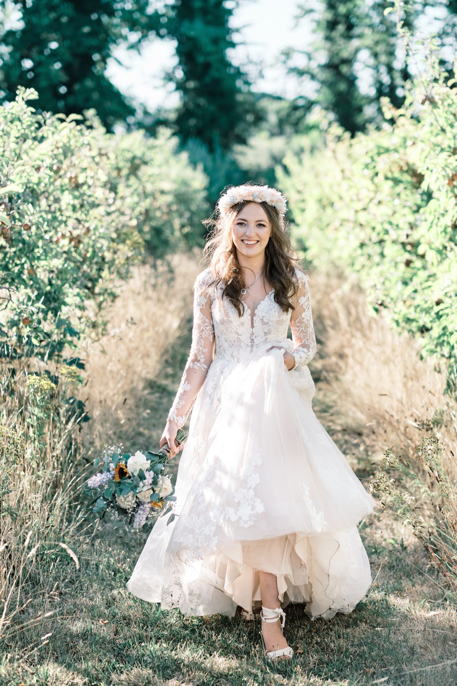 A bride in a flowing lace dress walks through a sunlit meadow holding a blue and white bouquet.