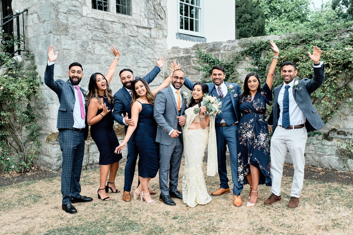 Wedding party celebrates outdoors in front of stone building with joyful poses and gestures.