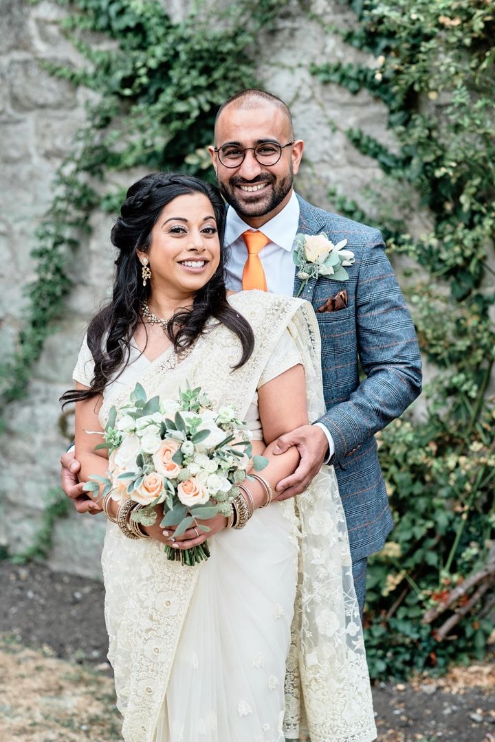 Newlyweds pose together against ivy-covered stone wall, bride holding peach and white bouquet.