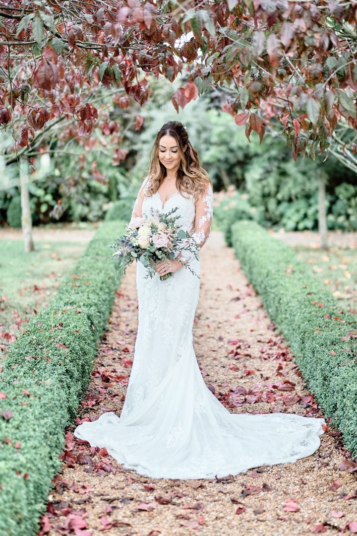 Bride in lace gown poses on garden path lined with manicured hedges.