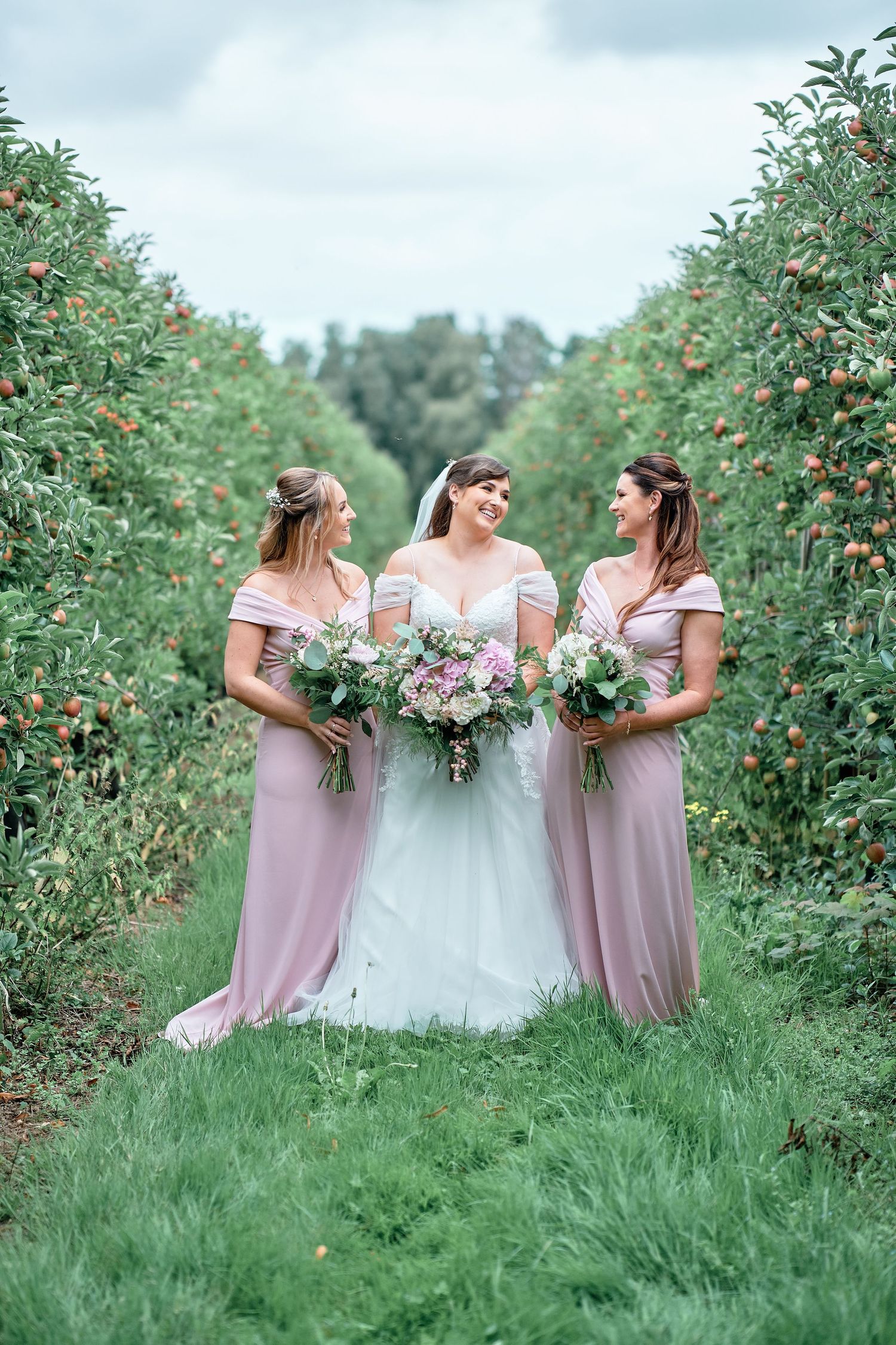 Wedding party in white and pink dresses poses in apple orchard with floral bouquets.